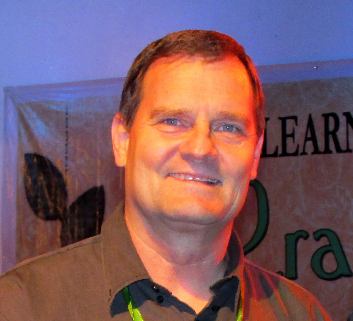 A smiling man with short brown hair and blue eyes, wearing a dark shirt and a green lanyard, standing in front of a banner with partially visible text and a leaf graphic.