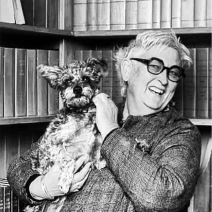A woman with glasses holding a small, curly-haired dog in front of a bookshelf.