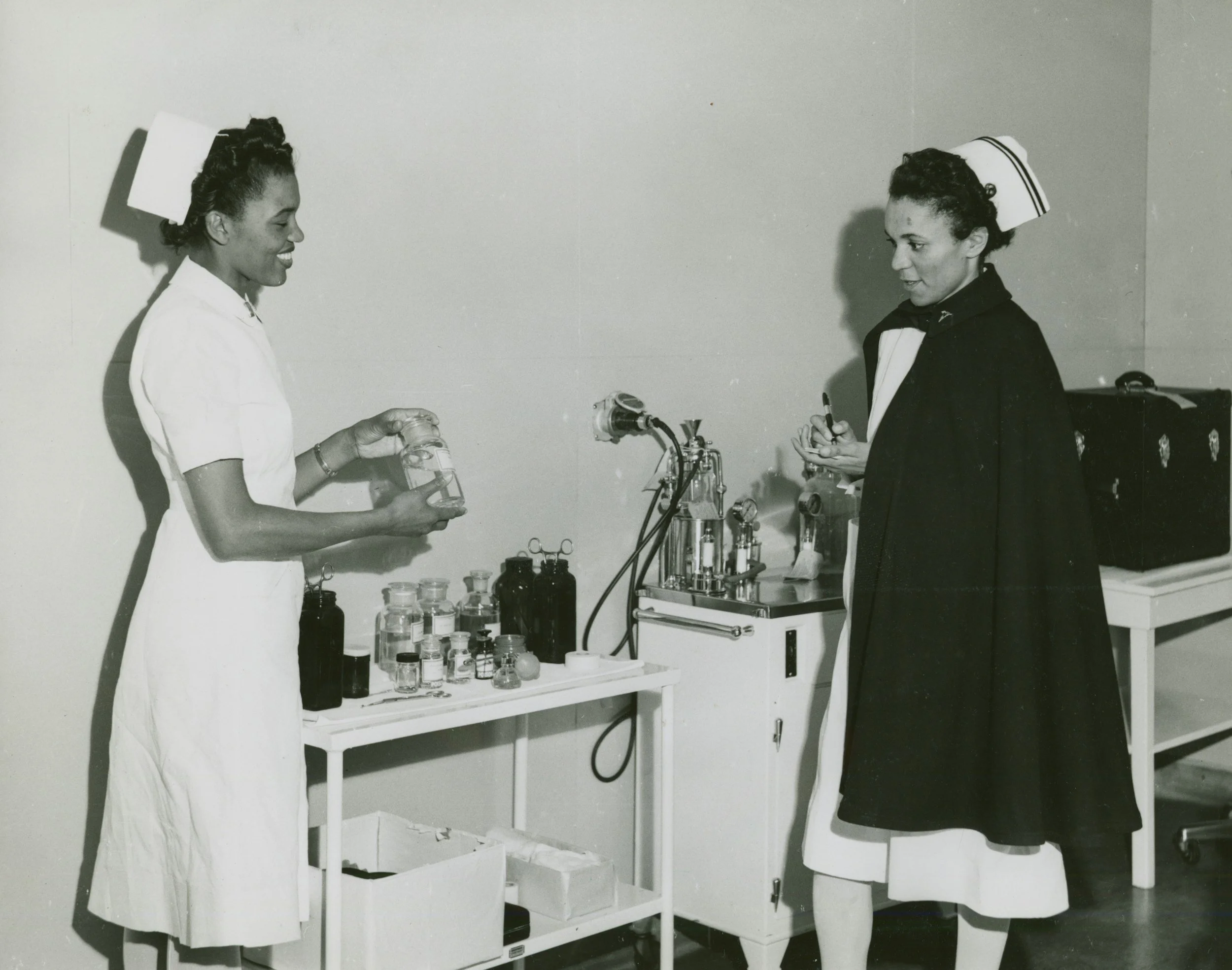 Black and white photo of two nurses in clinical uniforms standing in a medical laboratory or clinic, engaging in a discussion over scientific glassware and equipment on a table, with medical supplies in bottles and containers.