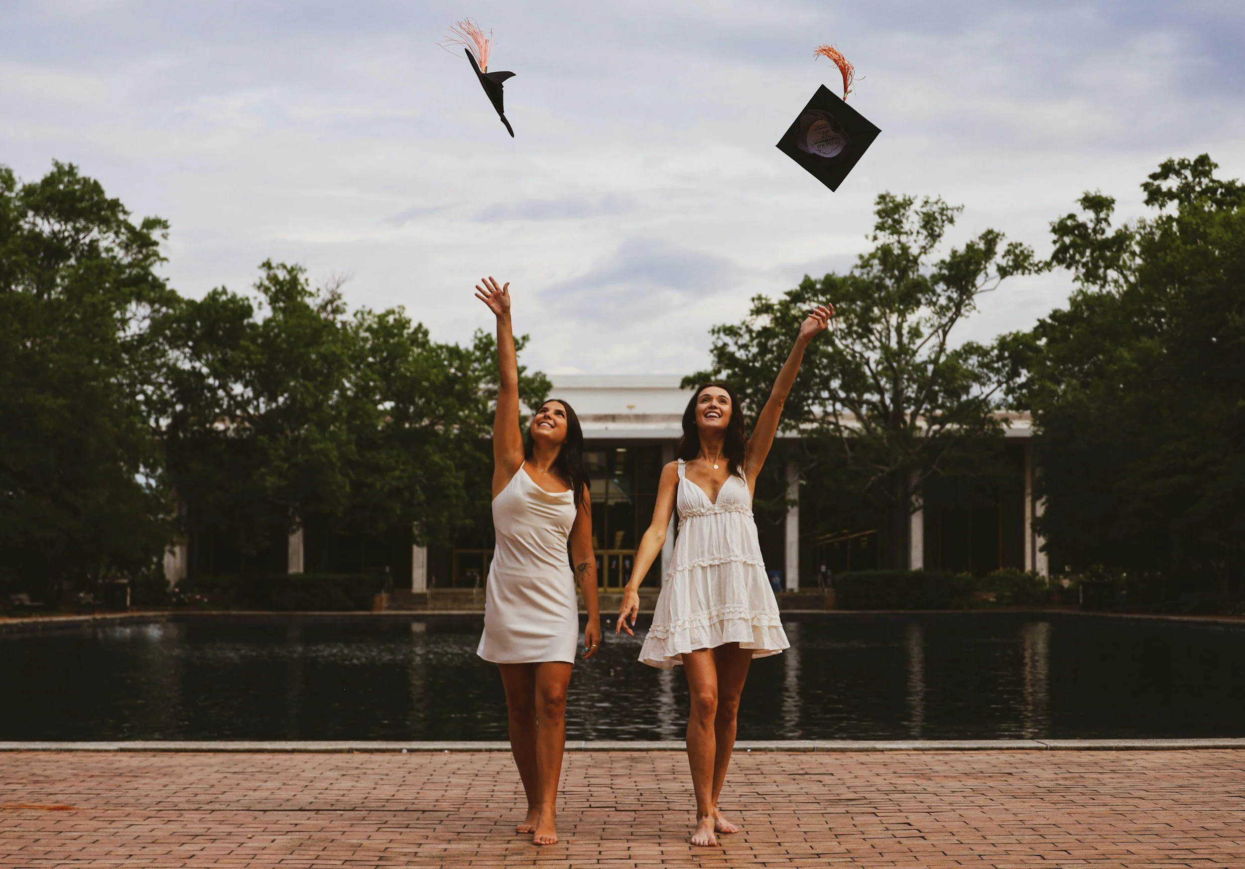 Two women in white dresses are standing on a brick path near a body of water, celebrating a graduation as they throw graduation caps into the air, with trees and a building in the background under a cloudy sky.