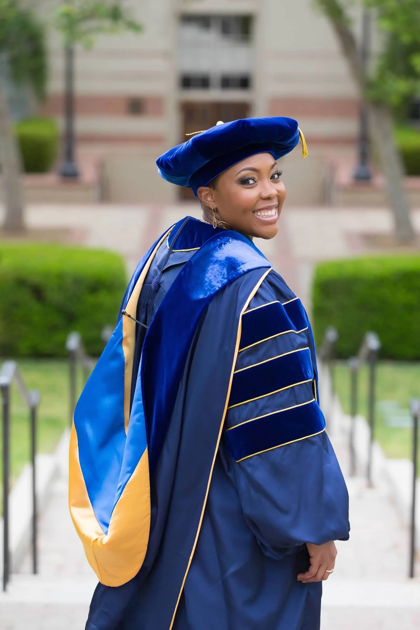 Woman in blue graduation gown and cap smiling outdoors in front of a building with trees and greenery.