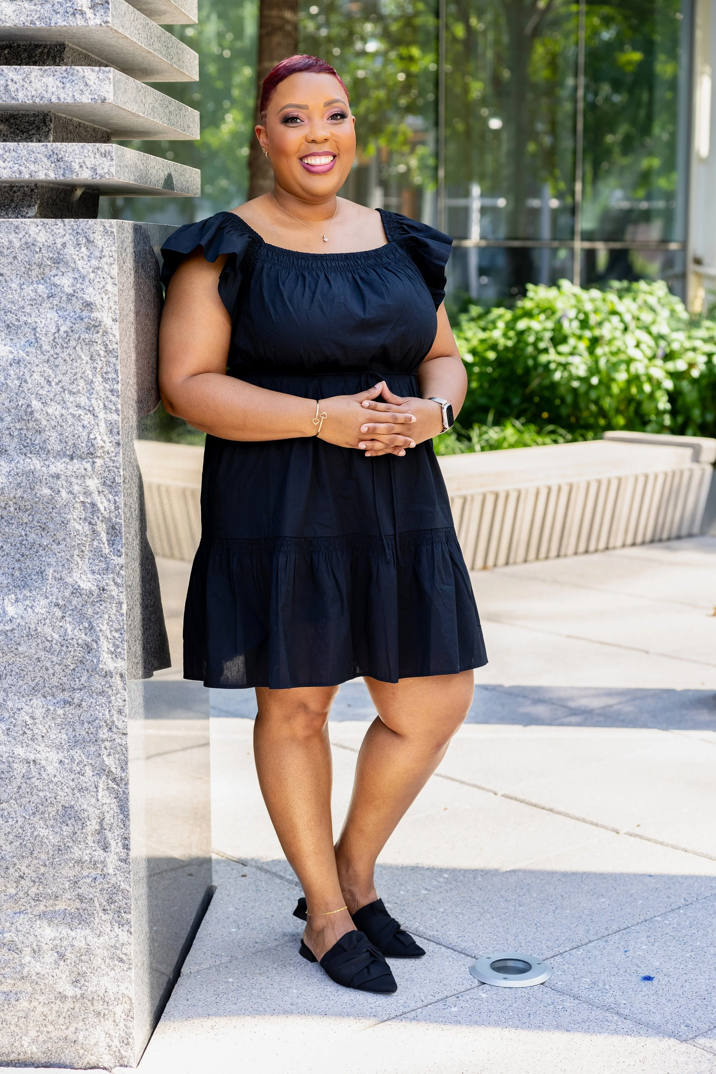 A woman wearing a black dress, black shoes, and jewelry, standing outdoors near a stone structure and a bench, smiling at the camera.