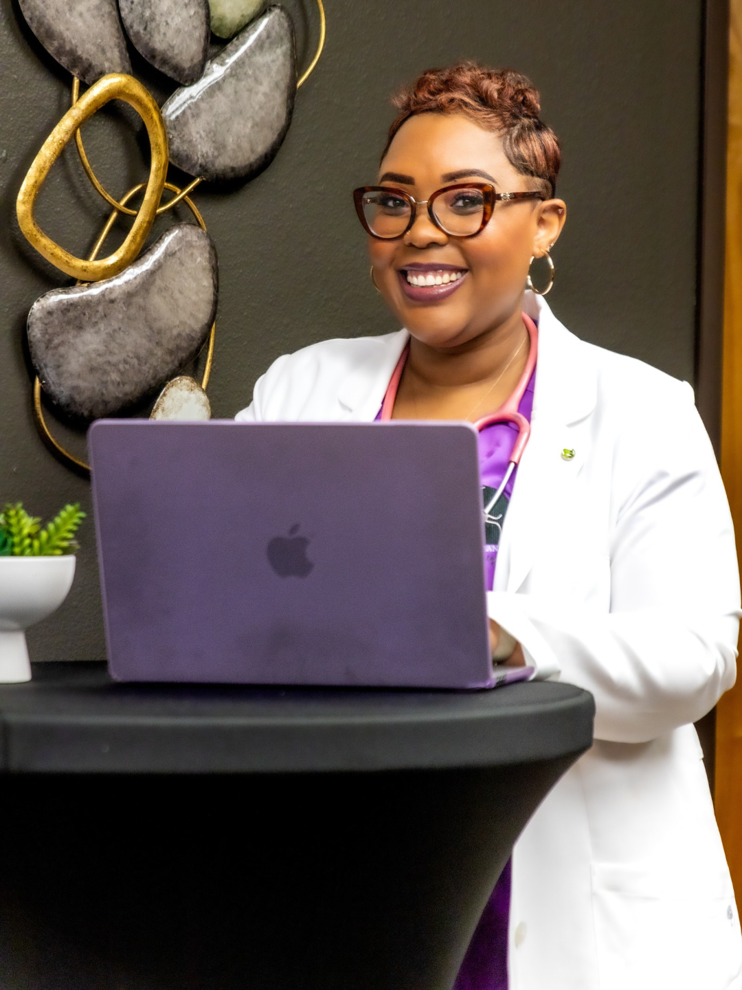 Dr. Tiffany Monique, a smiling nurse with short hair and glasses, sitting at a table with a purple laptop, wearing a white lab coat and purple scrubs, in a decorated room.