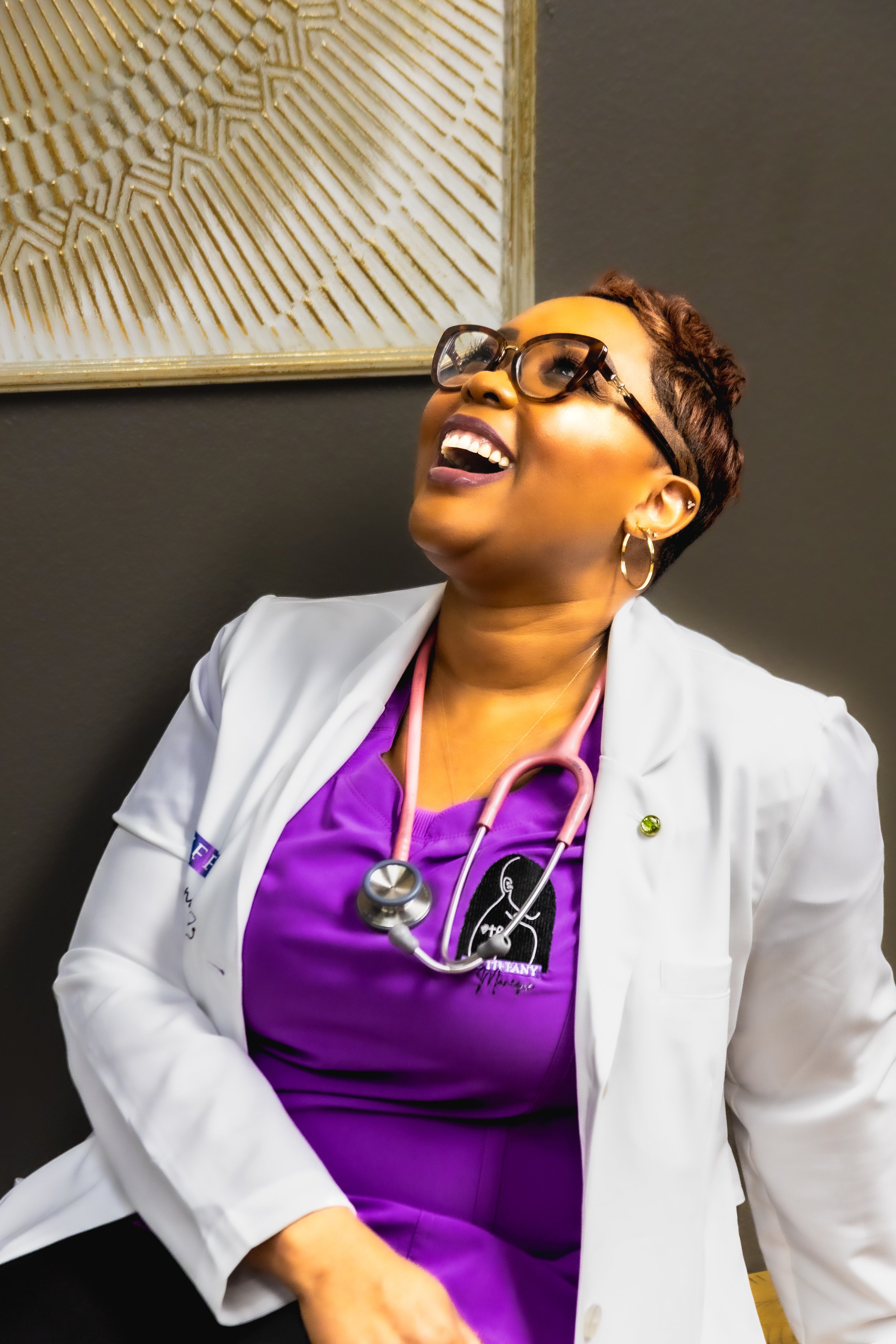A woman wearing glasses, a white medical coat, and purple scrubs with a stethoscope around her neck, smiling and looking up, against a dark wall with a decorative gold-striped artwork.