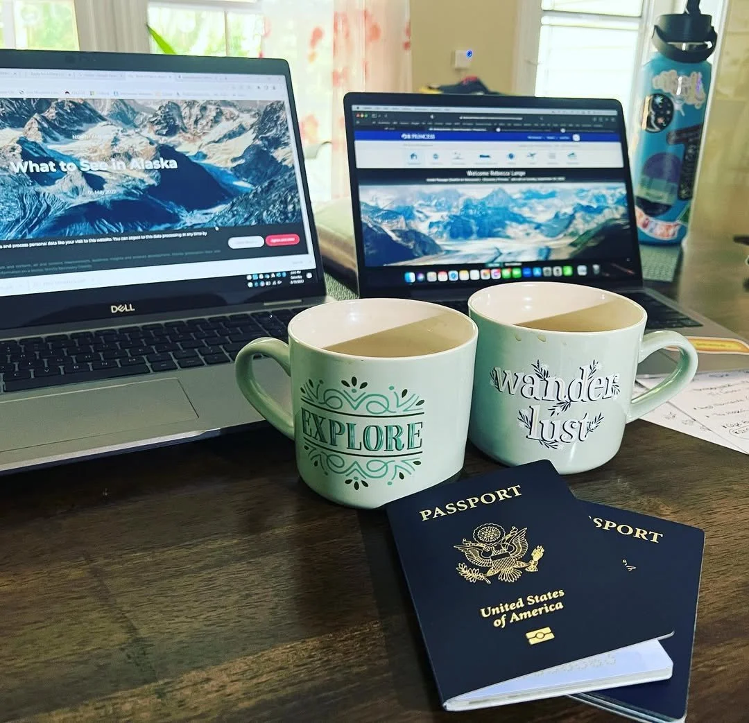 Two laptops on a wooden desk showing travel websites, two ceramic mugs with 'Explore' and 'Wander lust' written on them, two U.S. passports, a water bottle, and some paper notes.