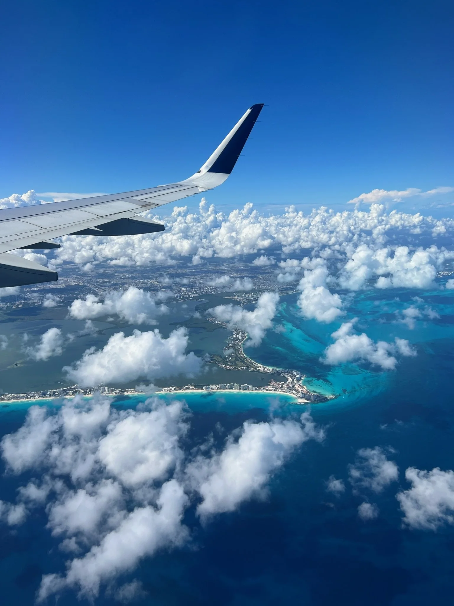 View from an airplane window showing airplane wing, clouds, turquoise water, and shoreline below.