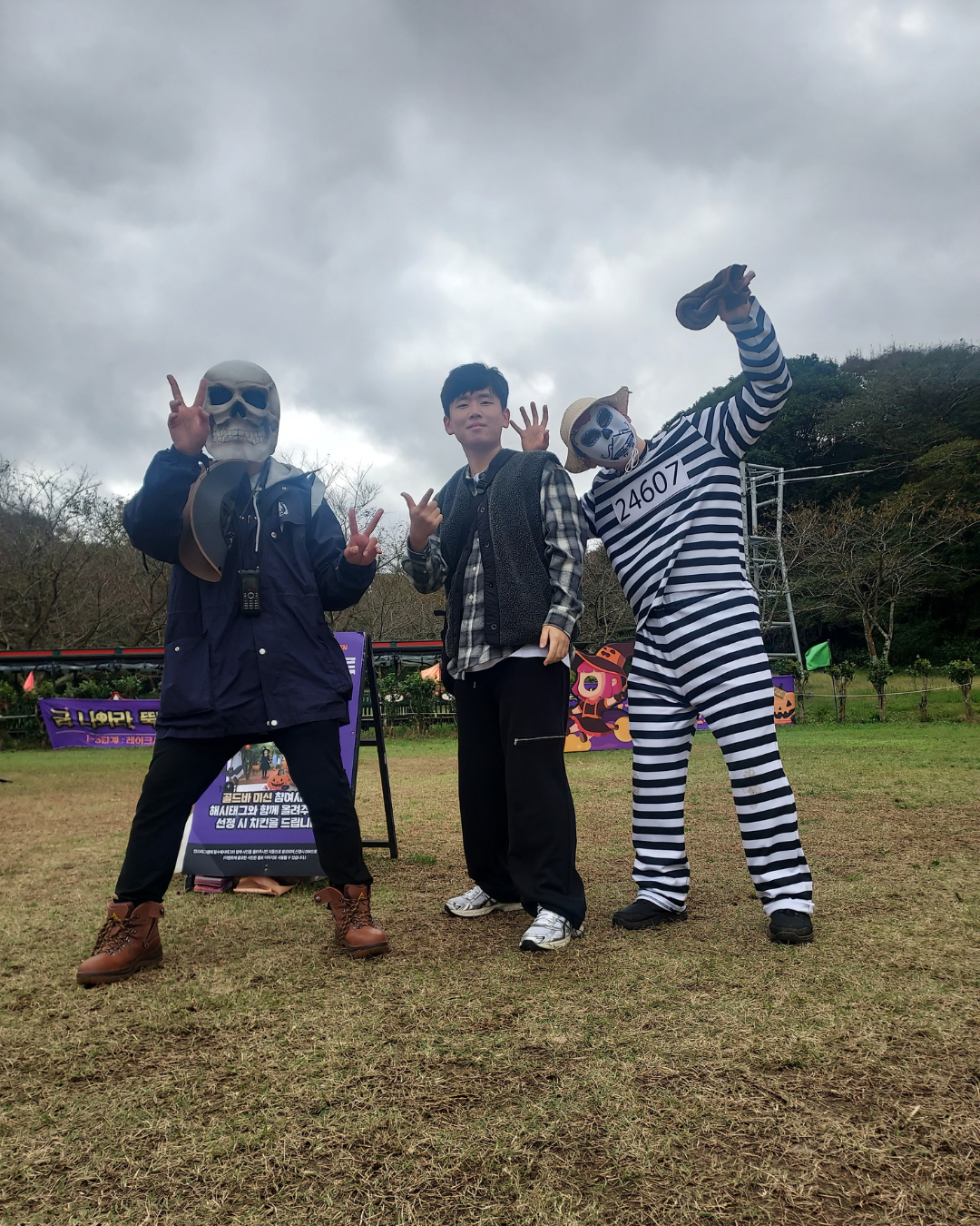 Three people in costume standing outdoors on a cloudy day, with one dressed as a skeleton, another in a striped prisoner costume with makeup, and a young man in casual clothing between them.
