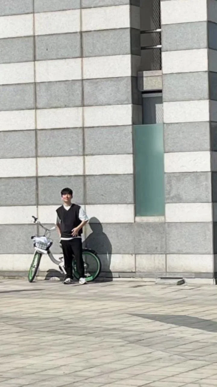 A young man standing next to a bicycle with a basket in front of a gray, striped building wall.