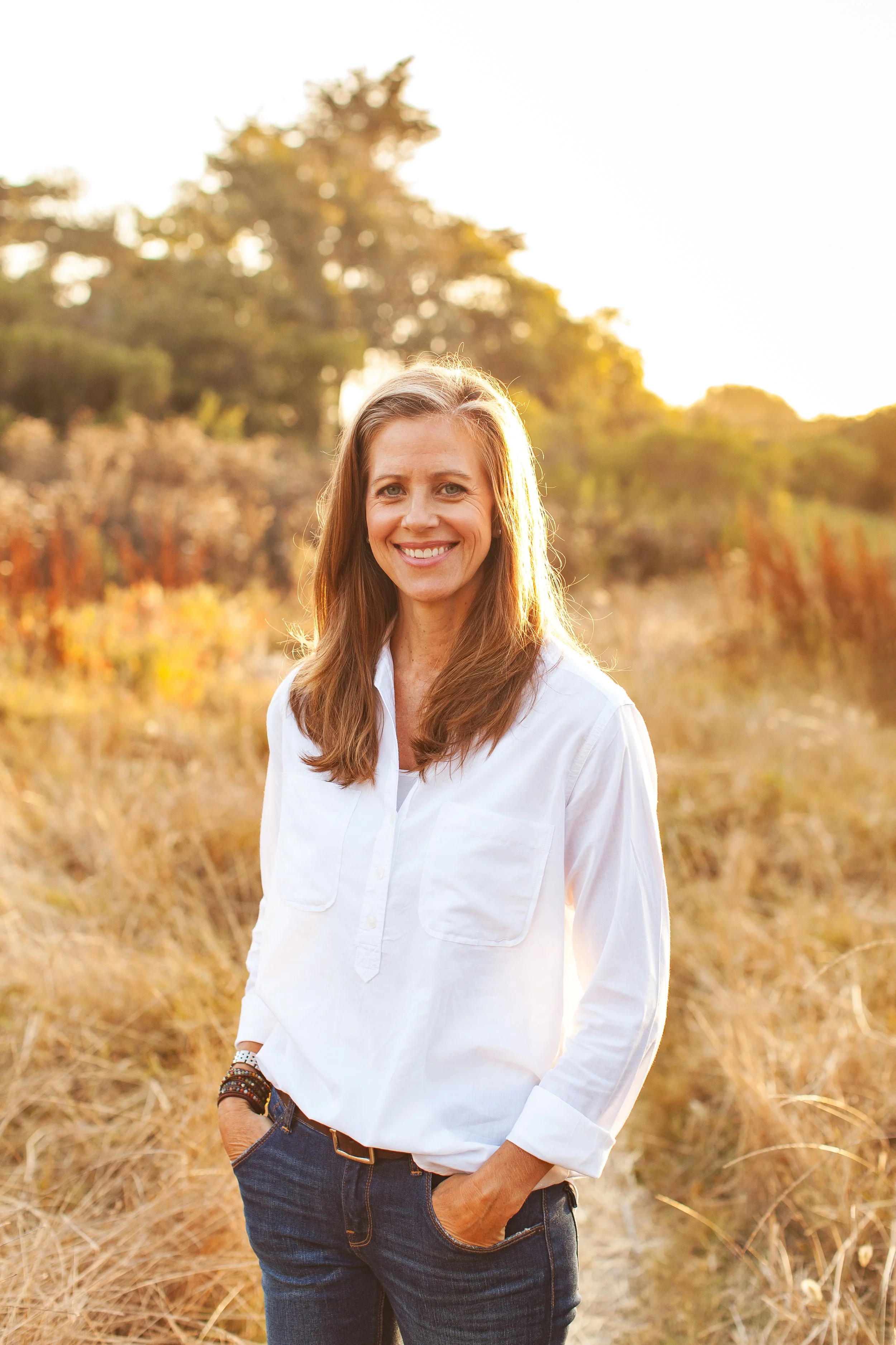 A woman standing in a field of dry grass during sunset, smiling and wearing a white shirt and blue jeans.