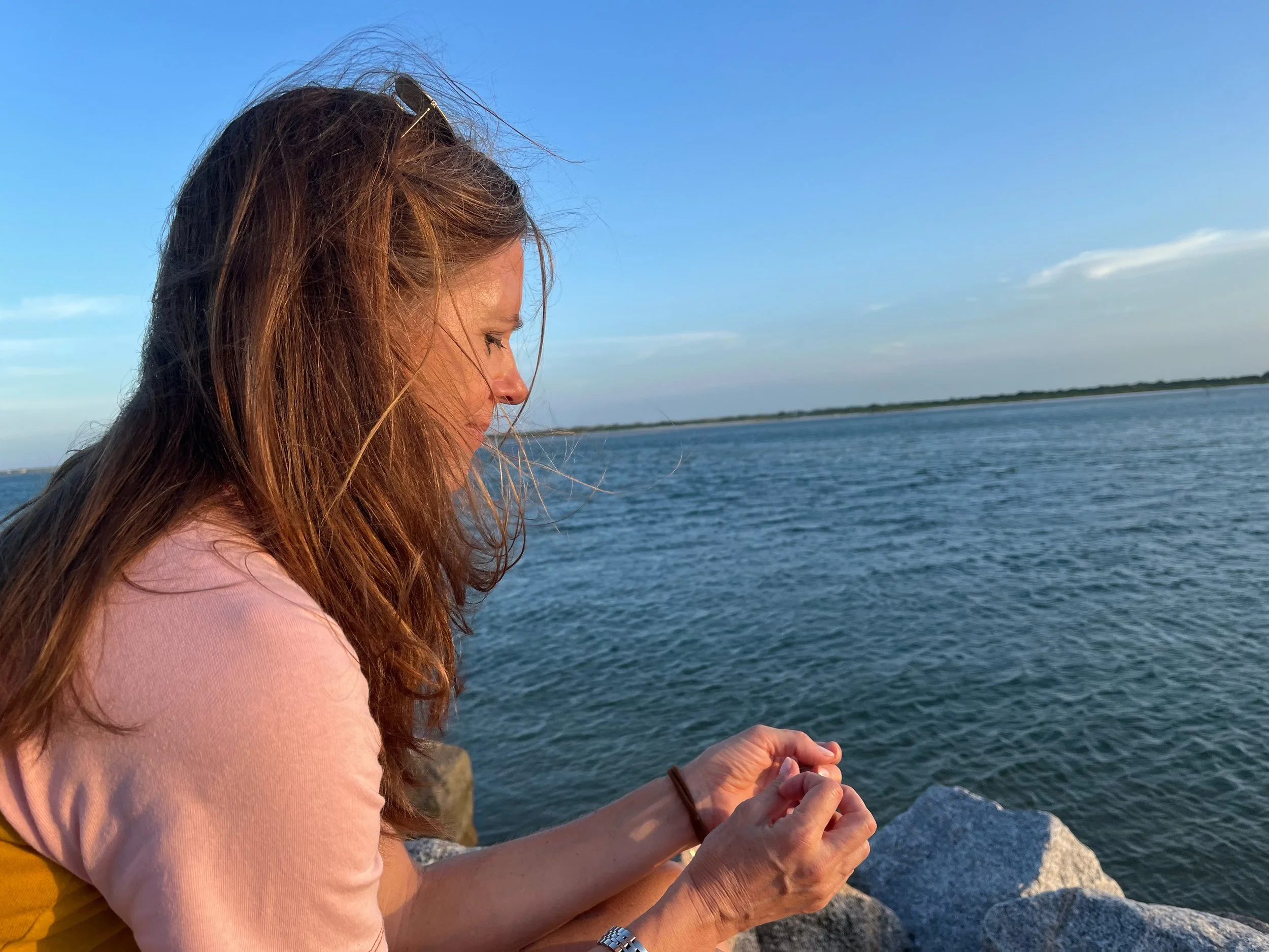 A woman with long, wavy hair and sunglasses on her head stands near the water, holding her hands together, overlooking the ocean or a large body of water with land in the background. She is wearing a pink long-sleeve shirt.