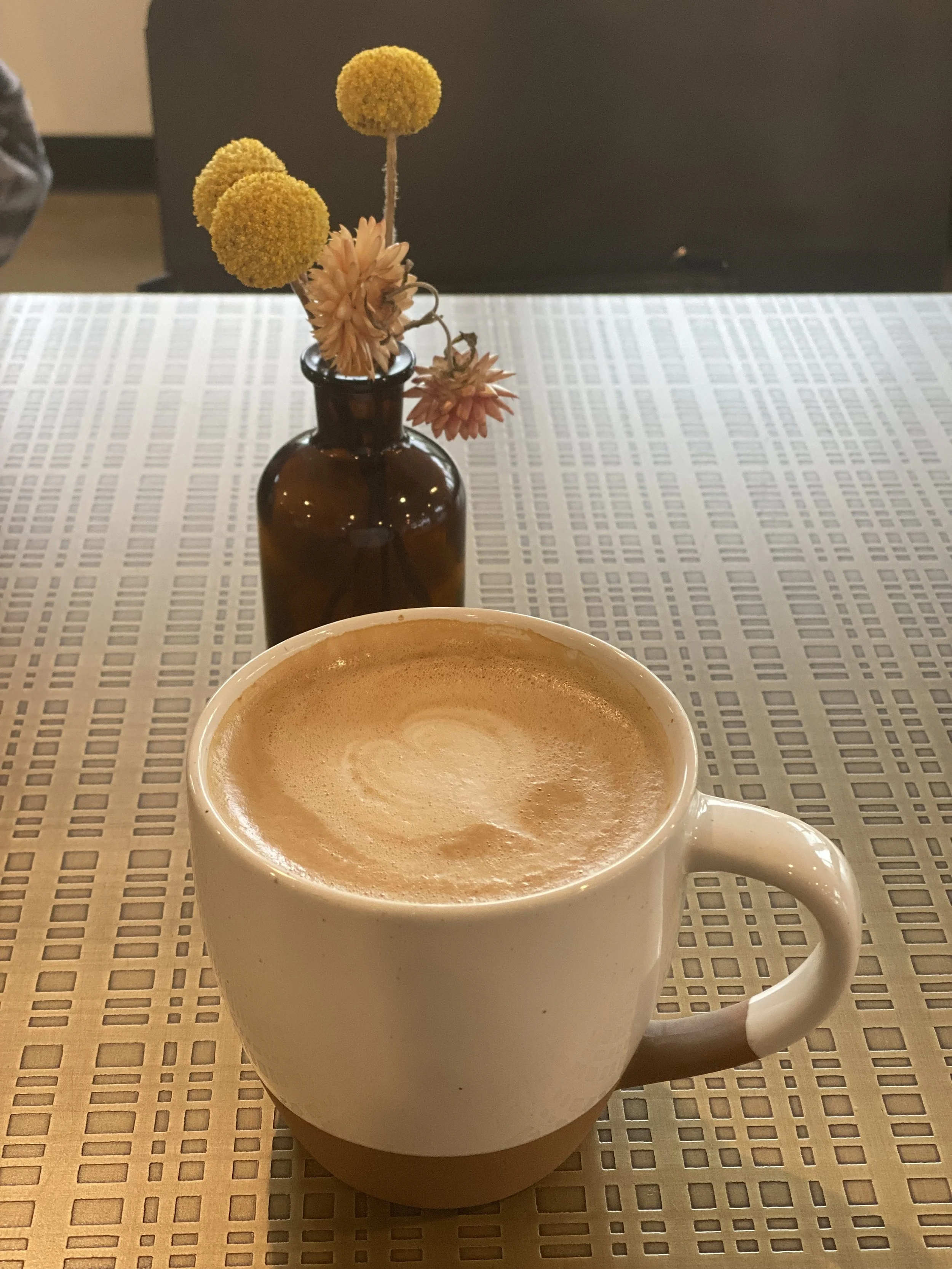 A white ceramic mug filled with frothy coffee on a table, with a brown glass vase holding dried flowers in the background.