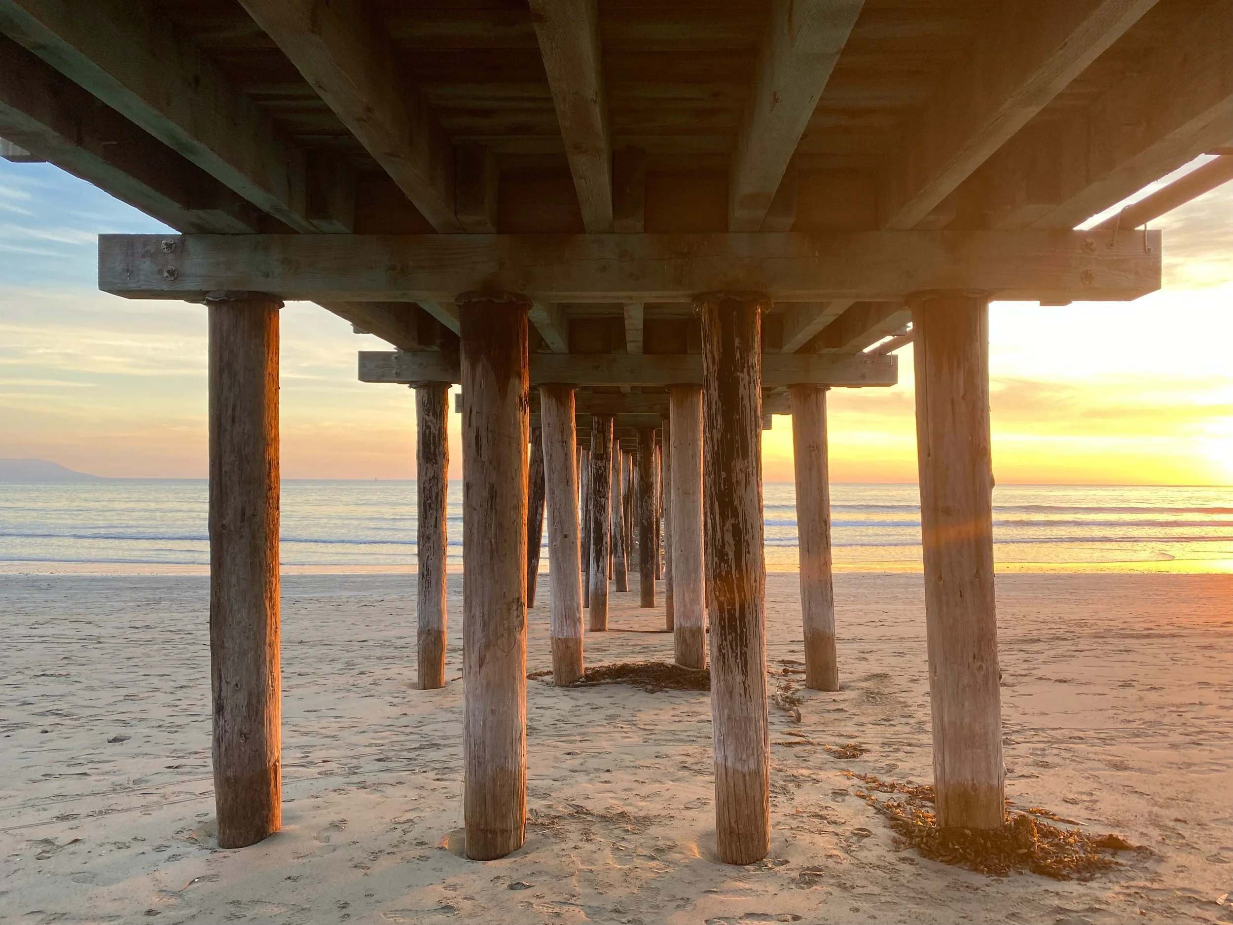 Underneath a wooden pier at sunset, with the pier's wood and supporting beams visible, overlooking a calm ocean and sandy beach.