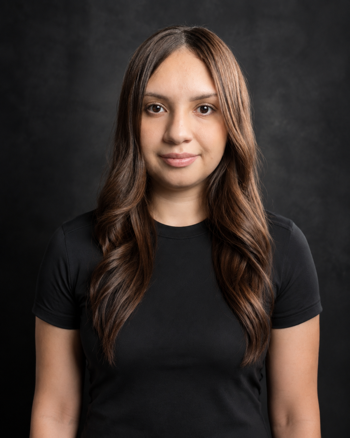 Portrait of a young woman with long, wavy brown hair wearing a black T-shirt, standing against a dark background.