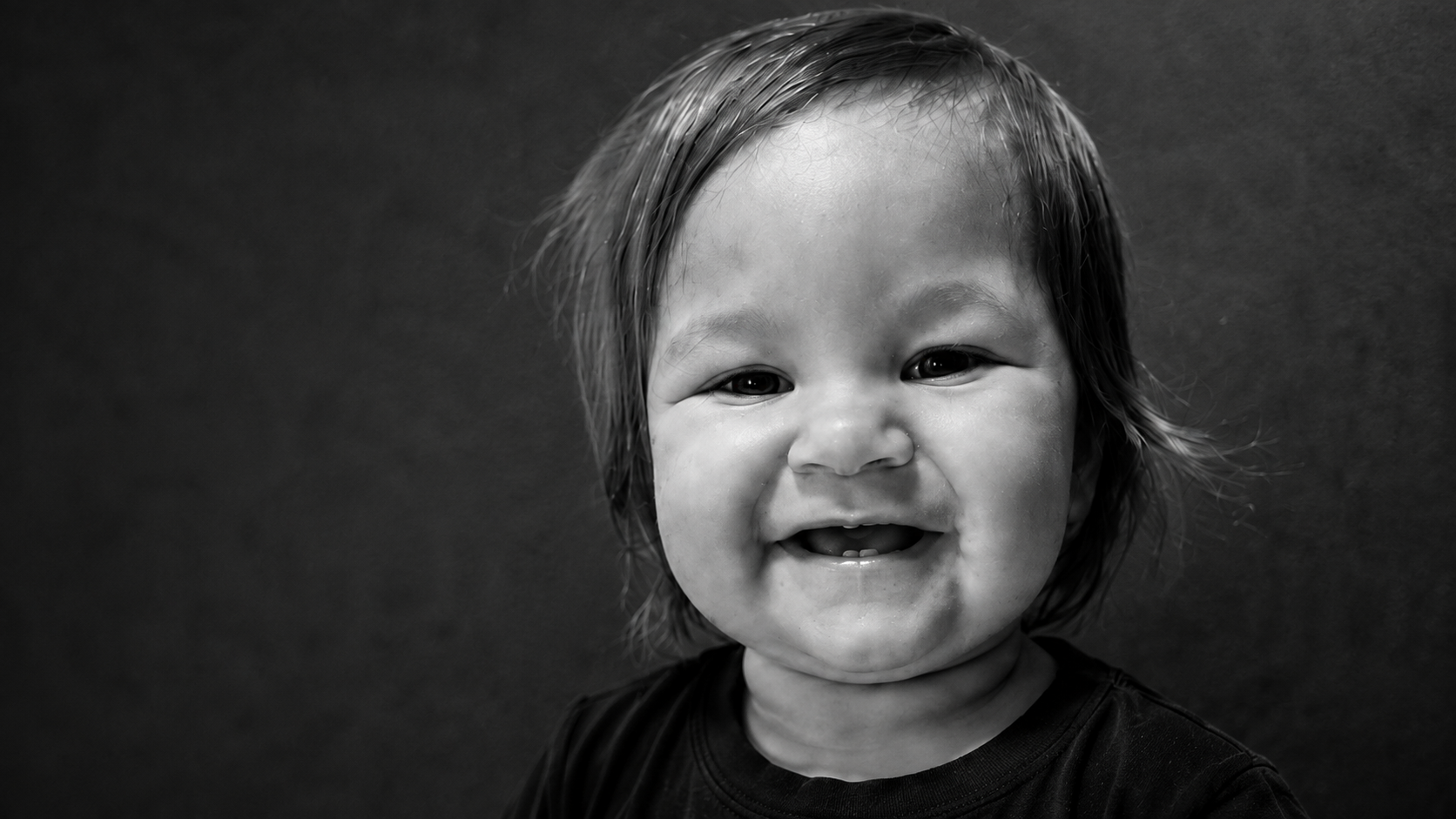 Black and white photo of a smiling young child with short, wet hair and a black shirt against a dark background.