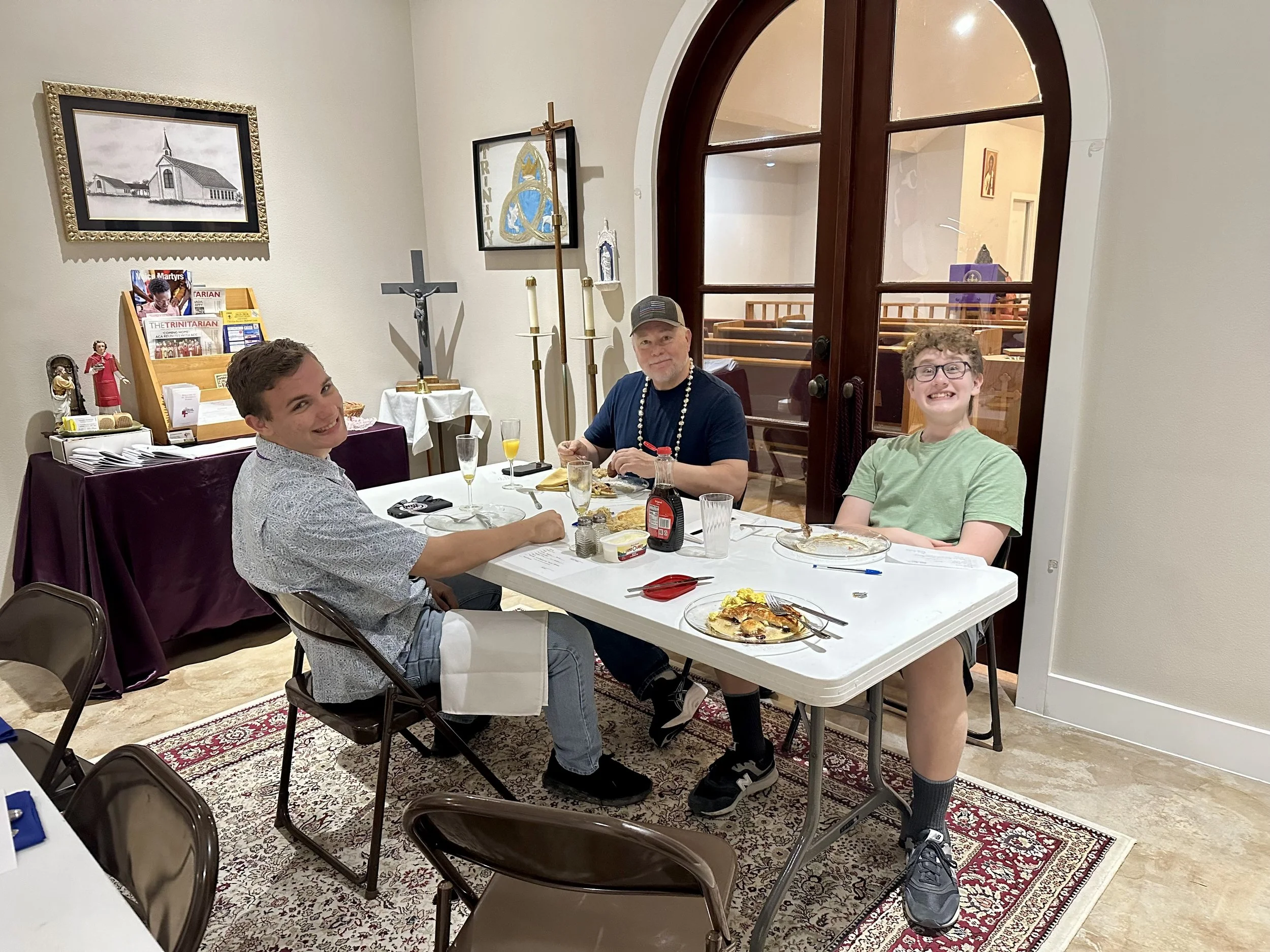 Three men celebrating around a table with food and drinks in a room decorated with religious pictures and symbols.
