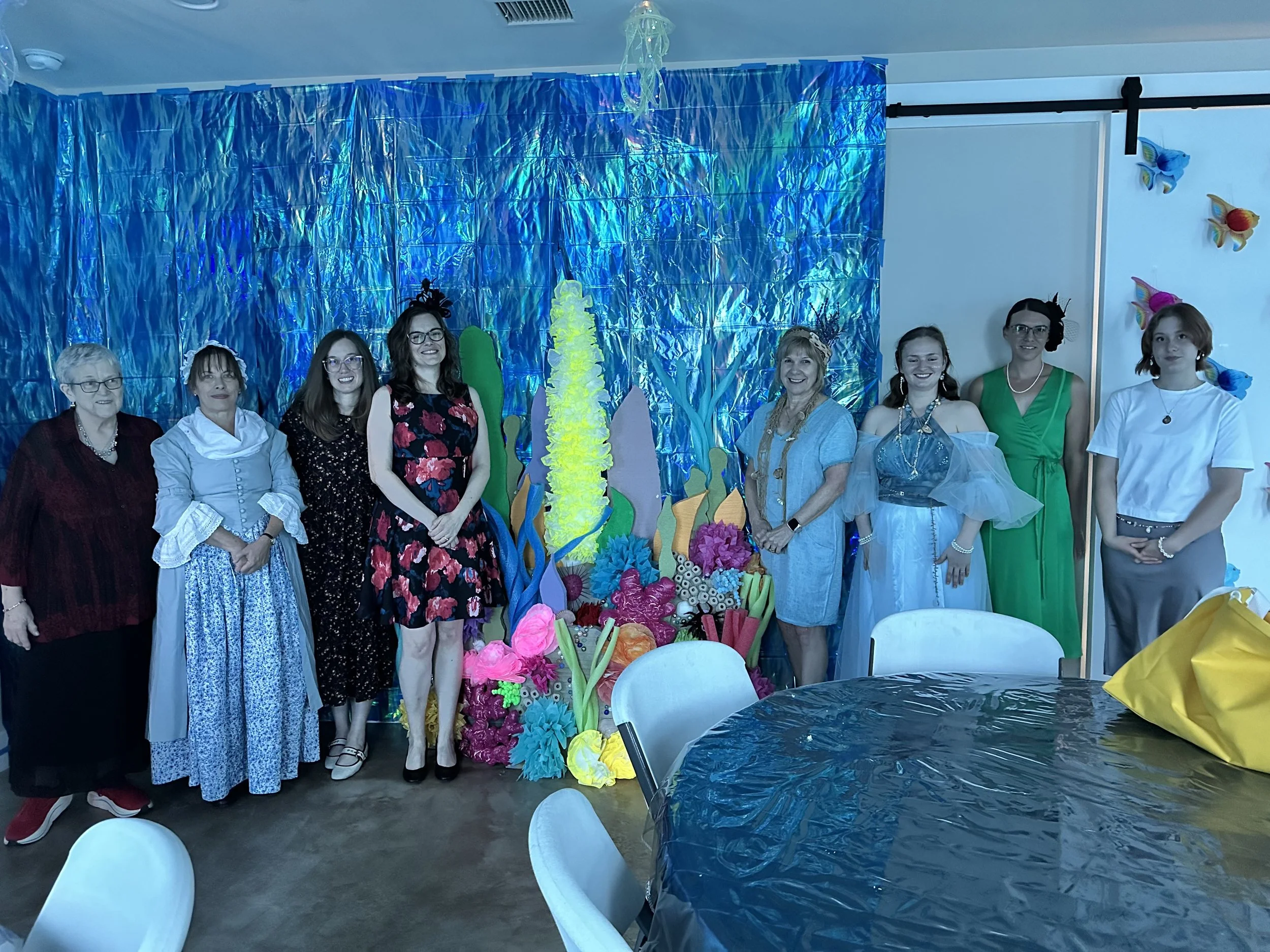 A group of eight women standing together at a decorated event with a blue foil curtain backdrop, colorful paper flower decorations, and tables covered with dark blue tablecloths in the foreground.