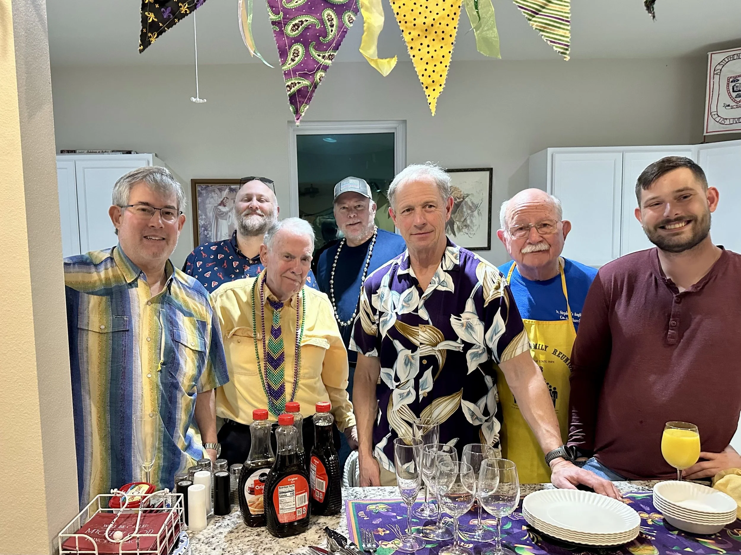 A group of eight men is gathered in a kitchen, celebrating with drinks, wearing colorful party accessories and Hawaiian shirts. There are festive decorations hanging from the ceiling, including purple, black, yellow, and striped fabric pennants. The countertop has bottles of soda, glasses, paper plates, and a variety of beverages in bottles.