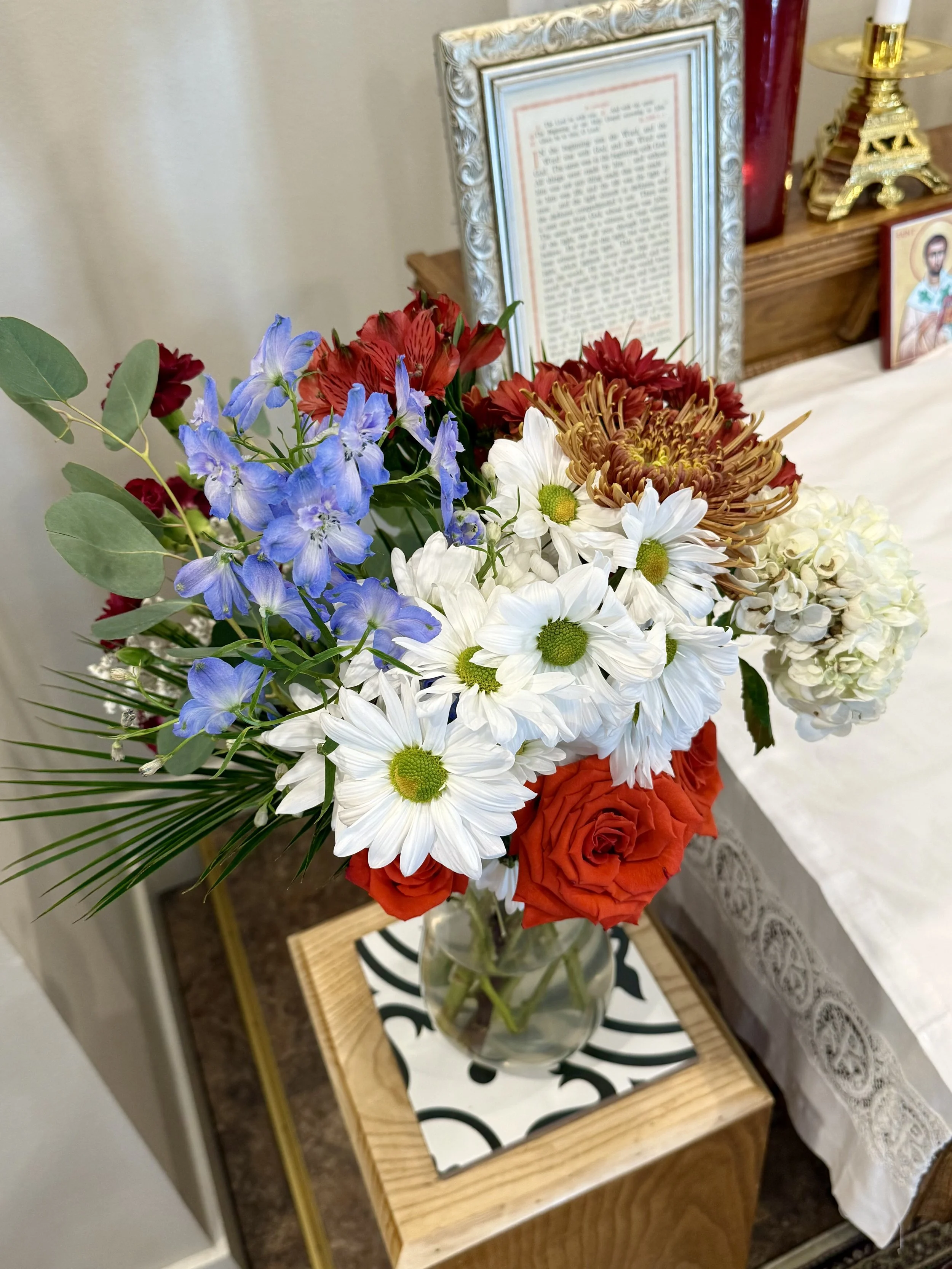 A bouquet of mixed flowers including white daisies, red roses, blue delphiniums, and other colorful flowers in a glass vase on a small wooden table.