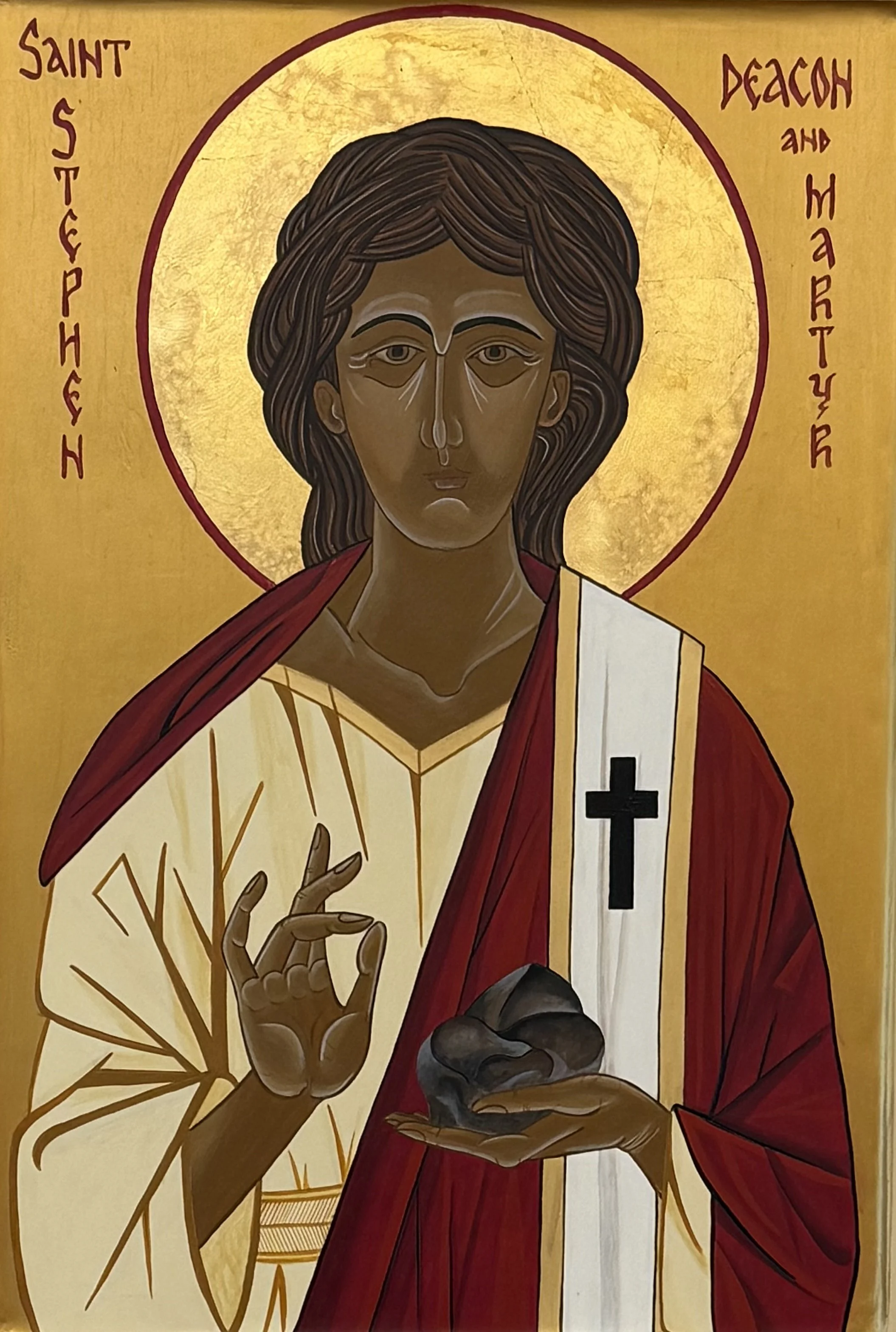 An icon depicting St. Stephen with a gold halo, wearing a red robe, making a gesture with his right hand and holding a dark object in his left hand, with an inscription near his head that reads 'Saint Stephen and Decon and Martyr' in stylized text.