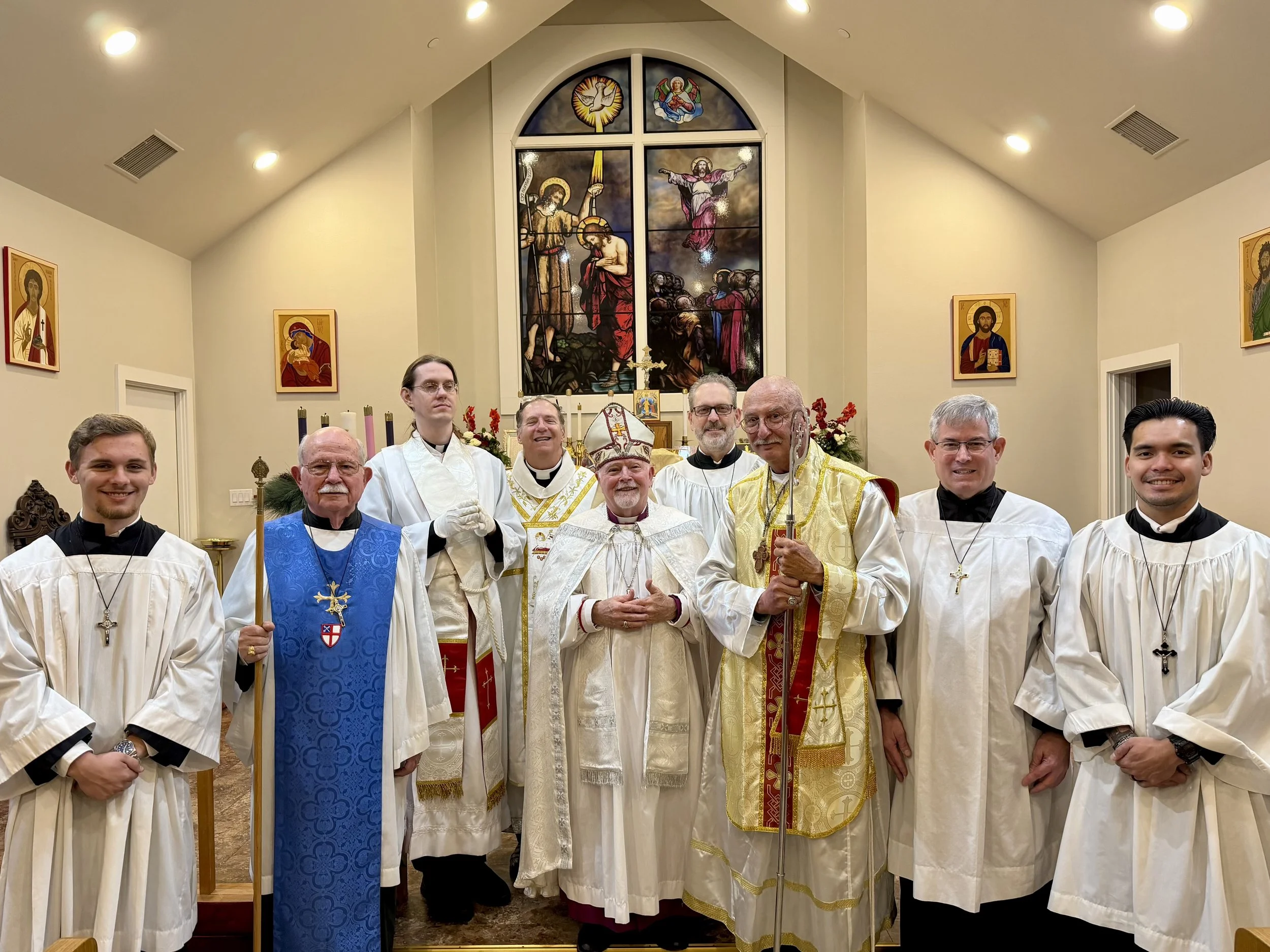 Group of clergy members, including bishops and priests, standing inside St. Stephens church with stained glass windows depicting religious scenes and icons in the background.