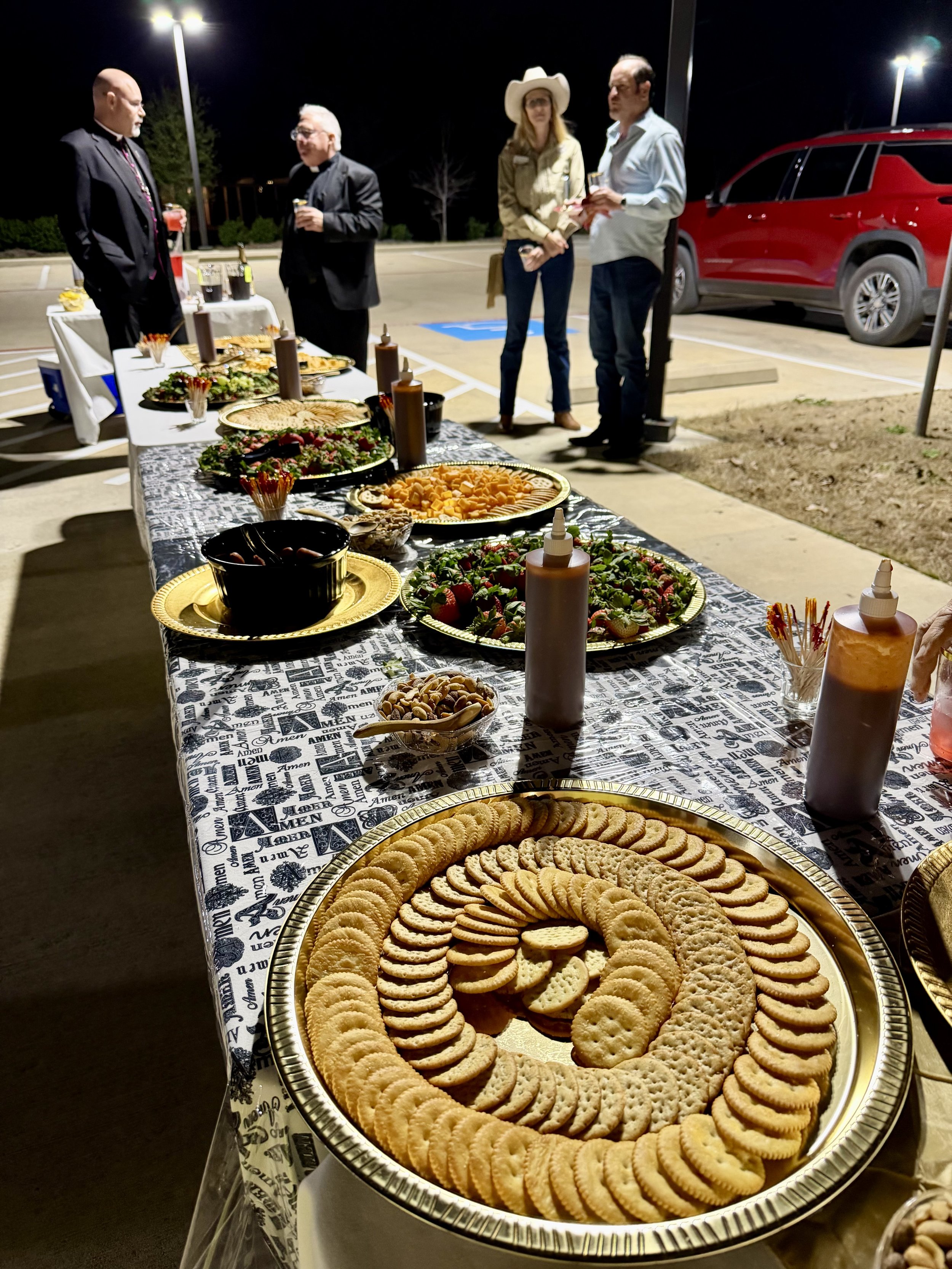 A long table covered with various plates of snacks, cookies, candies, and strawberry-covered desserts, set up outdoors at night with five people standing in the background