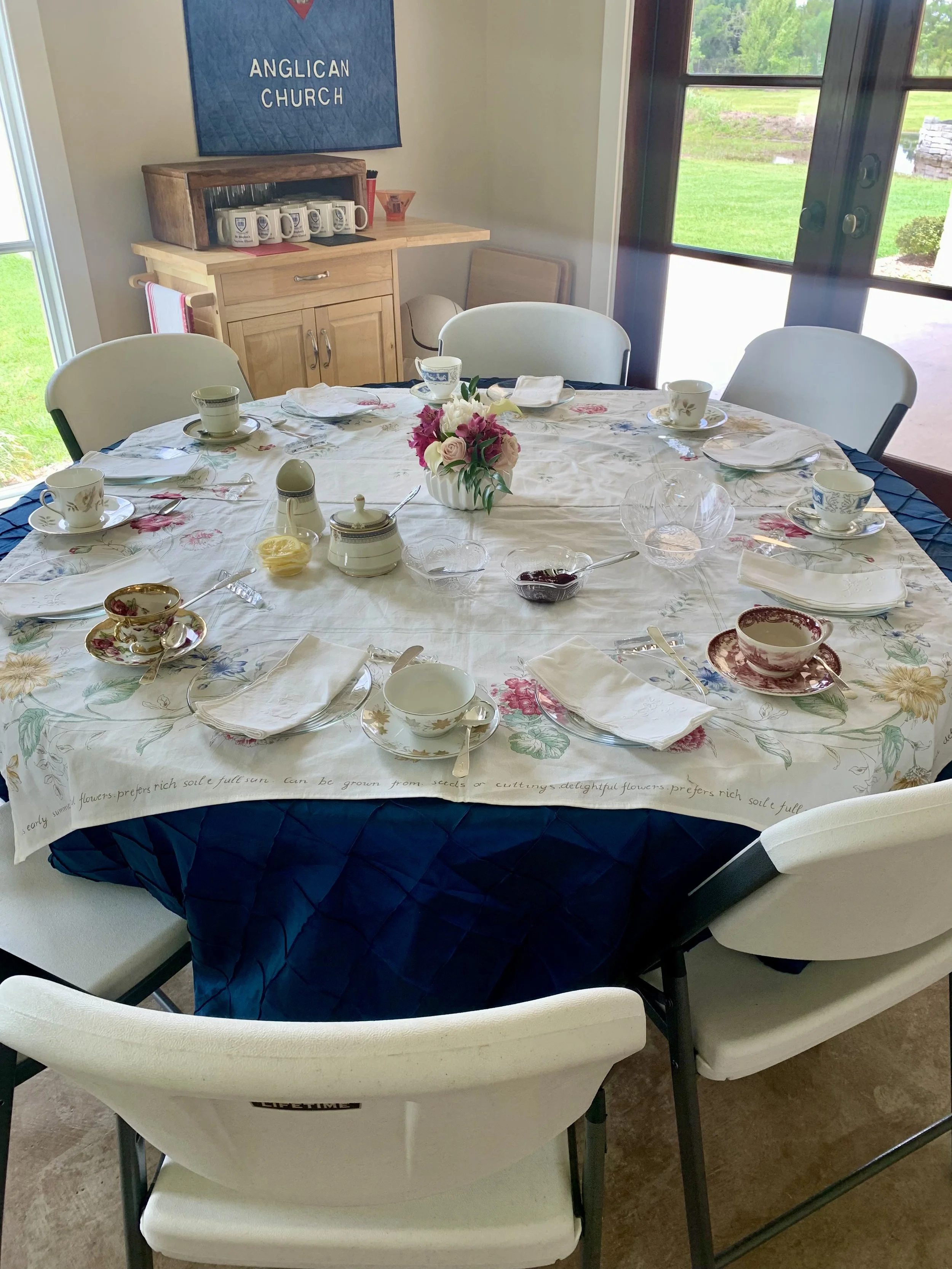 A dining table set for the Ladies Tea with a floral tablecloth, teacups, and bowls, with a centerpiece of fresh pink and white flowers, in the parish hall.