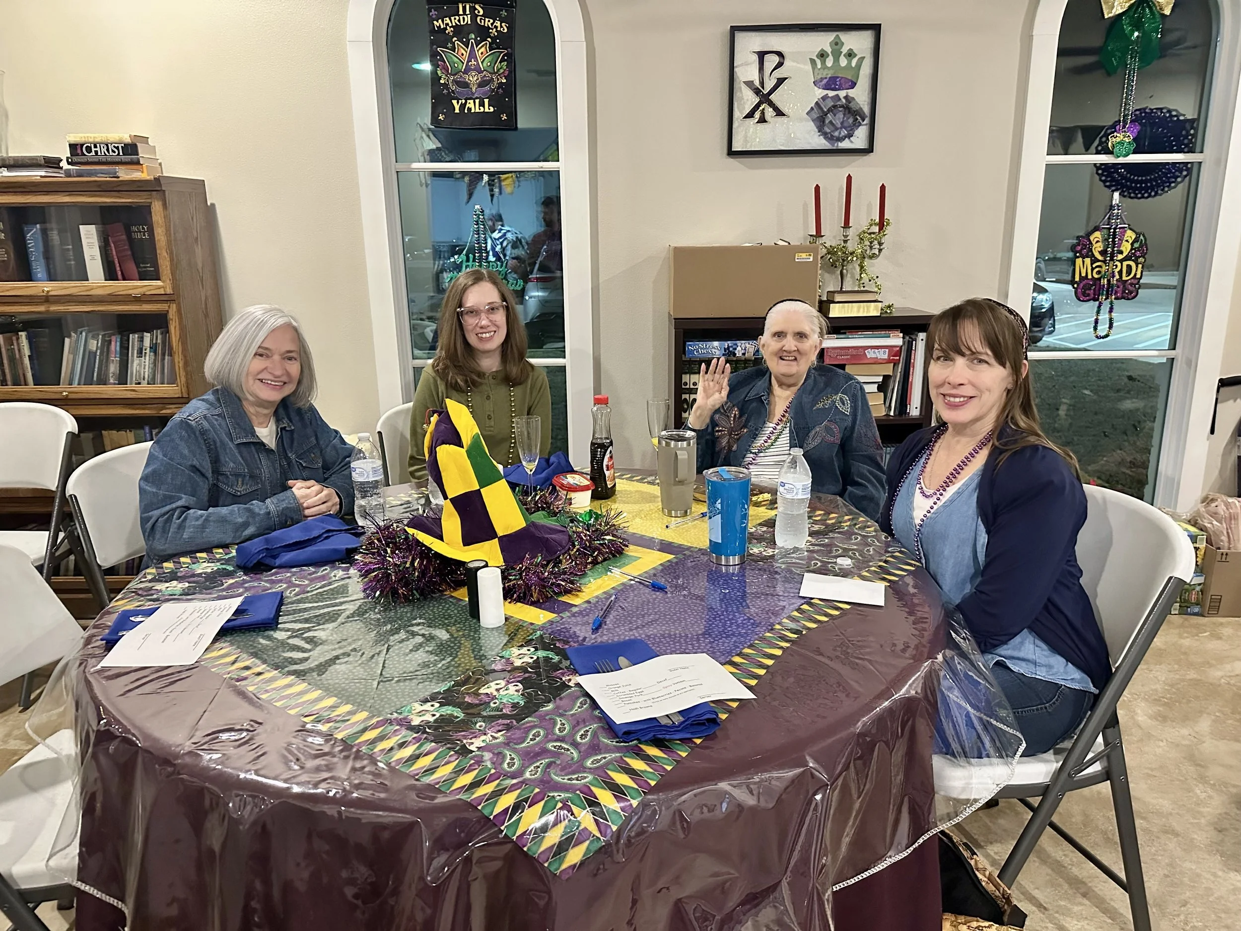 Four women sitting around a decorated table with Mardi Gras decorations, drinks, and printed menus in the parish hall.