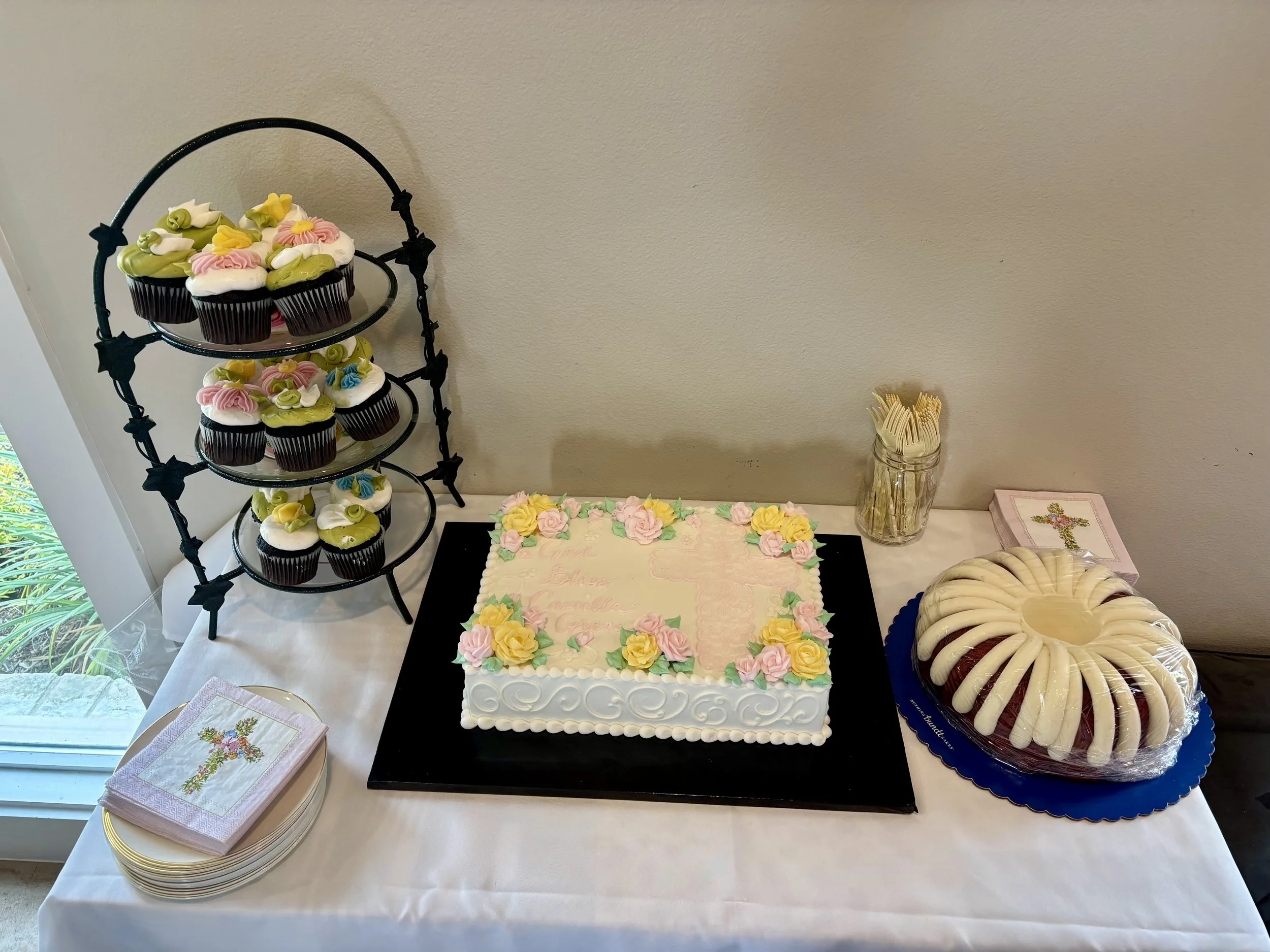 A decorated birthday cake with pink and yellow flowers on top, a cupcake stand with decorated cupcakes, a plate with cookies, and party napkins on a table near a window.