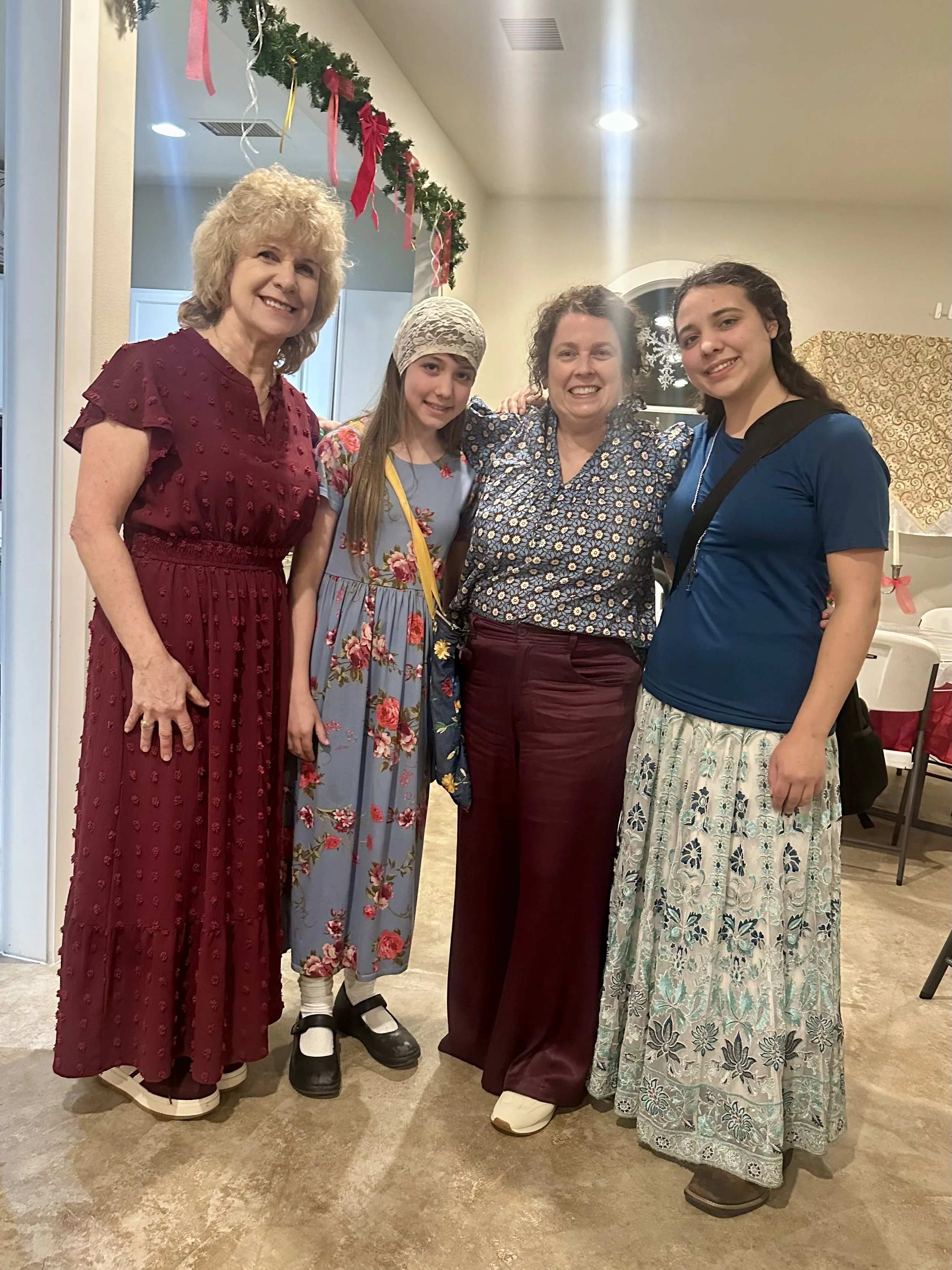 Four women standing together indoors during a holiday celebration, smiling, with Christmas decorations such as garland and bows hanging in the background.