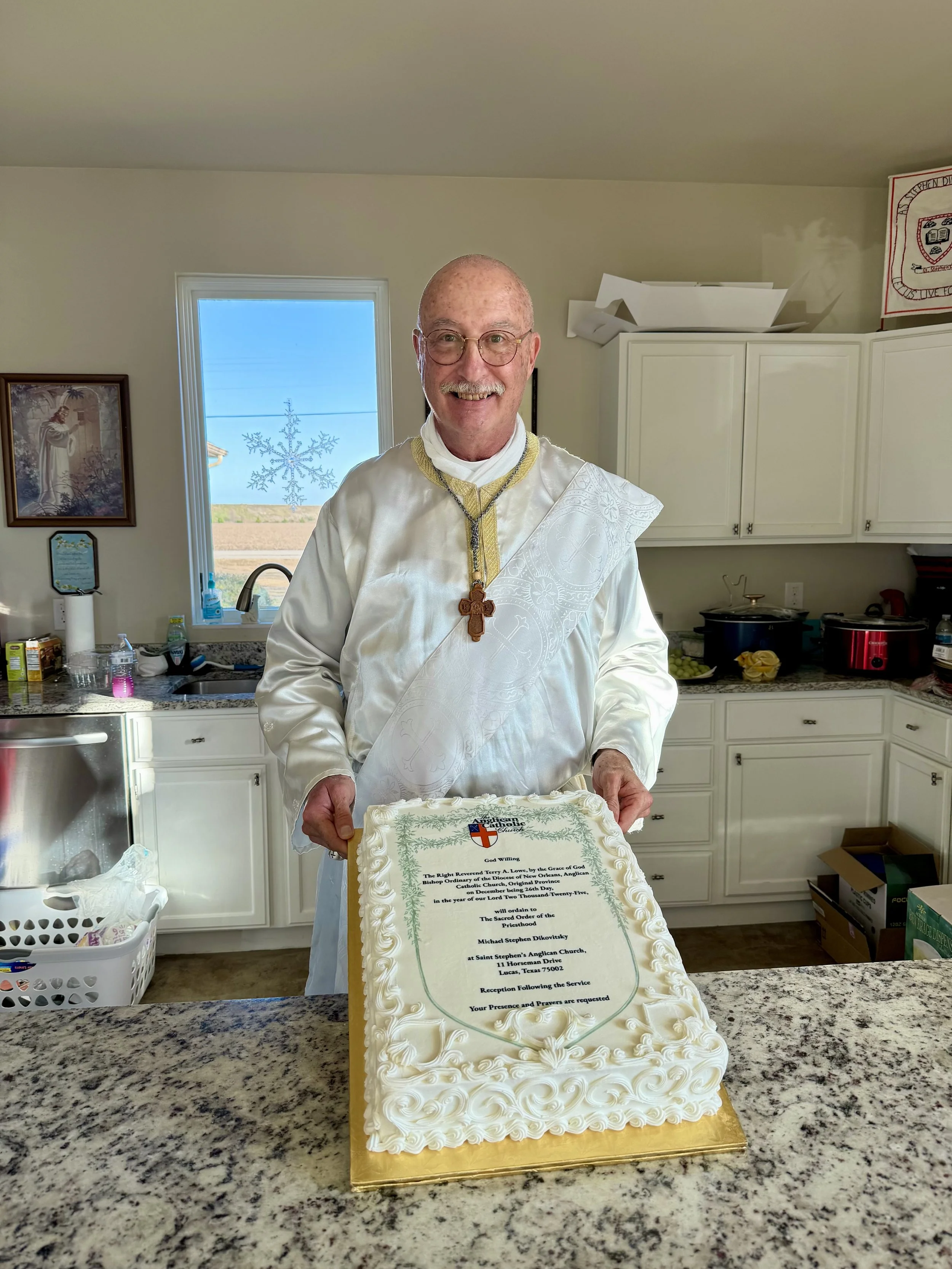 Fr. Micheal Dikovitsky holding an ornate cake with a religious message and a crest, standing in the kitchen.