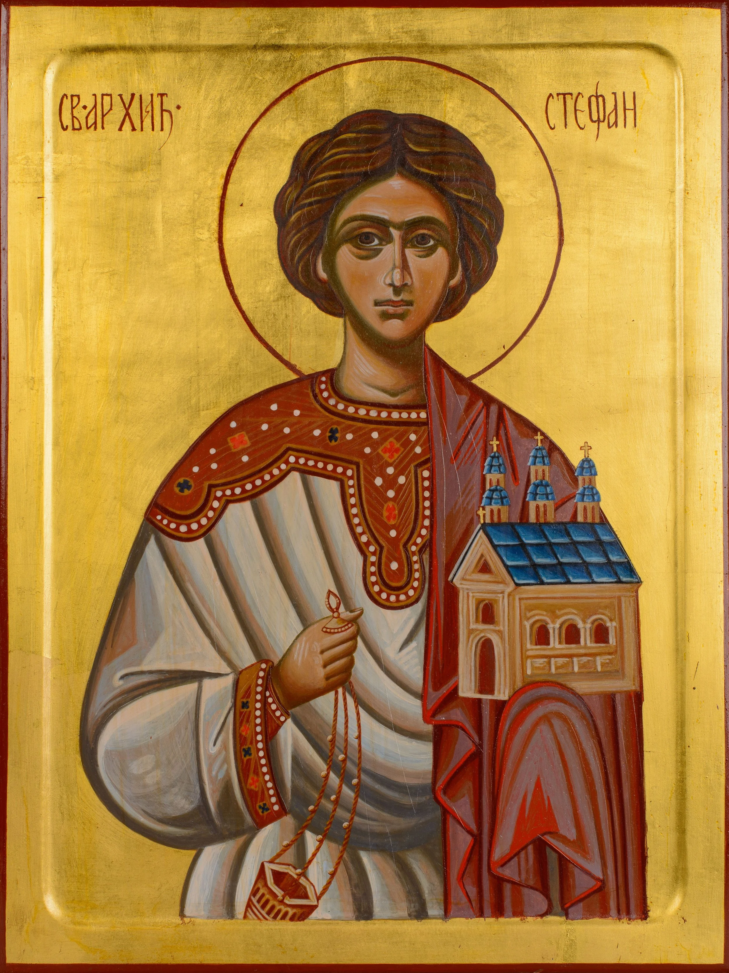 Icon of a saint with a golden background, wearing a white and red garment, holding a small church with blue domes in one hand and a rosary in the other.
