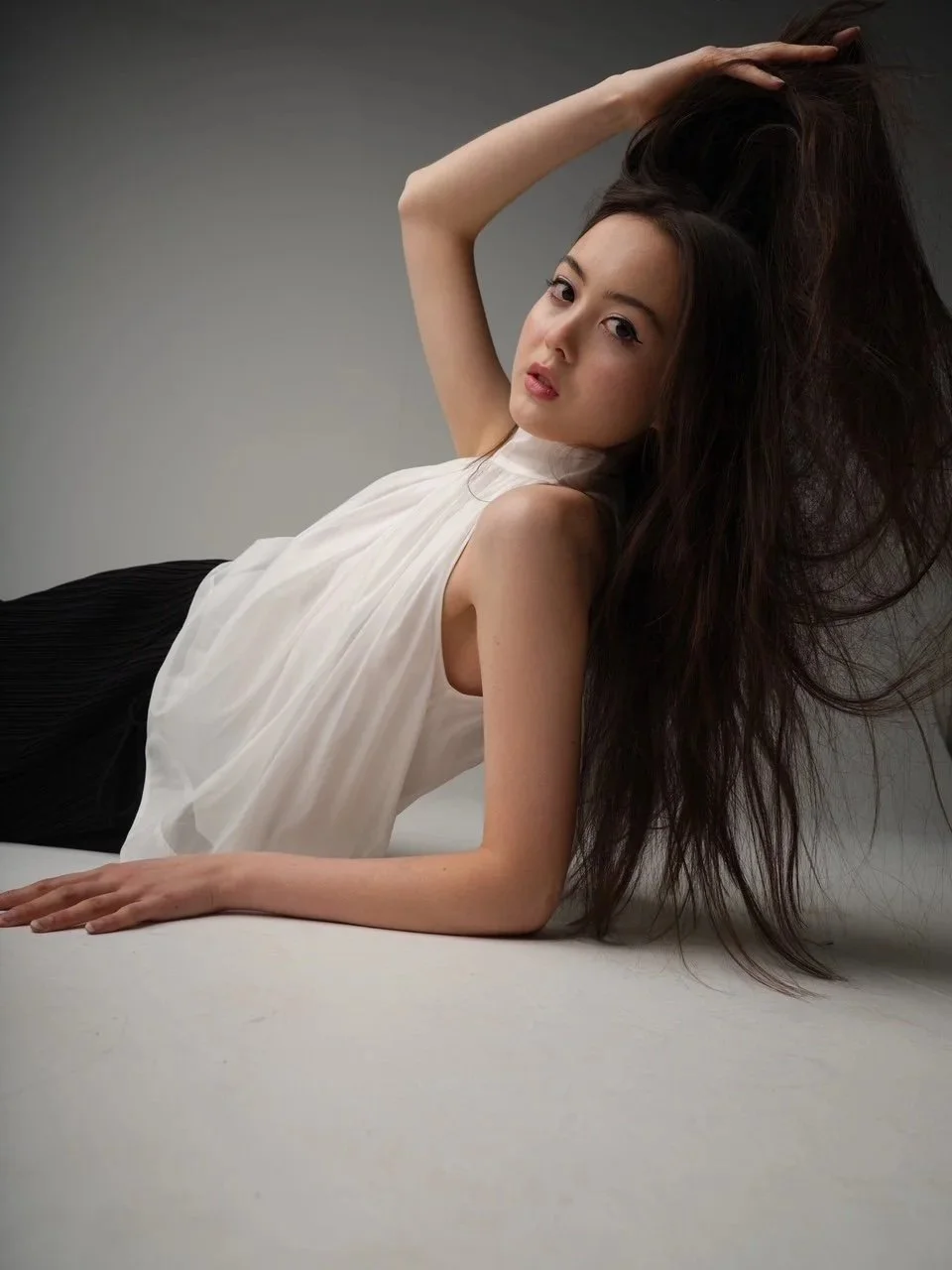 A young woman with long dark hair lying on the floor, wearing a sleeveless white top and black bottoms, with one arm raised and her hand in her hair, looking at the camera against a plain gray background.