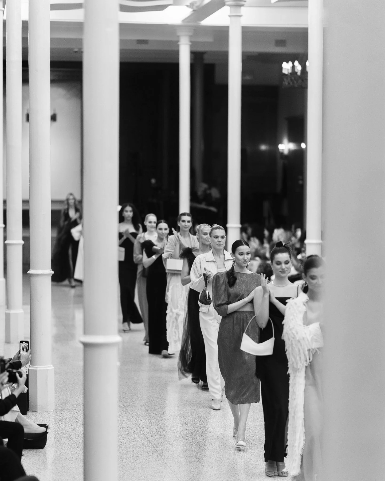 A group of women dressed in elegant clothing standing in a line inside a building with tall white columns.