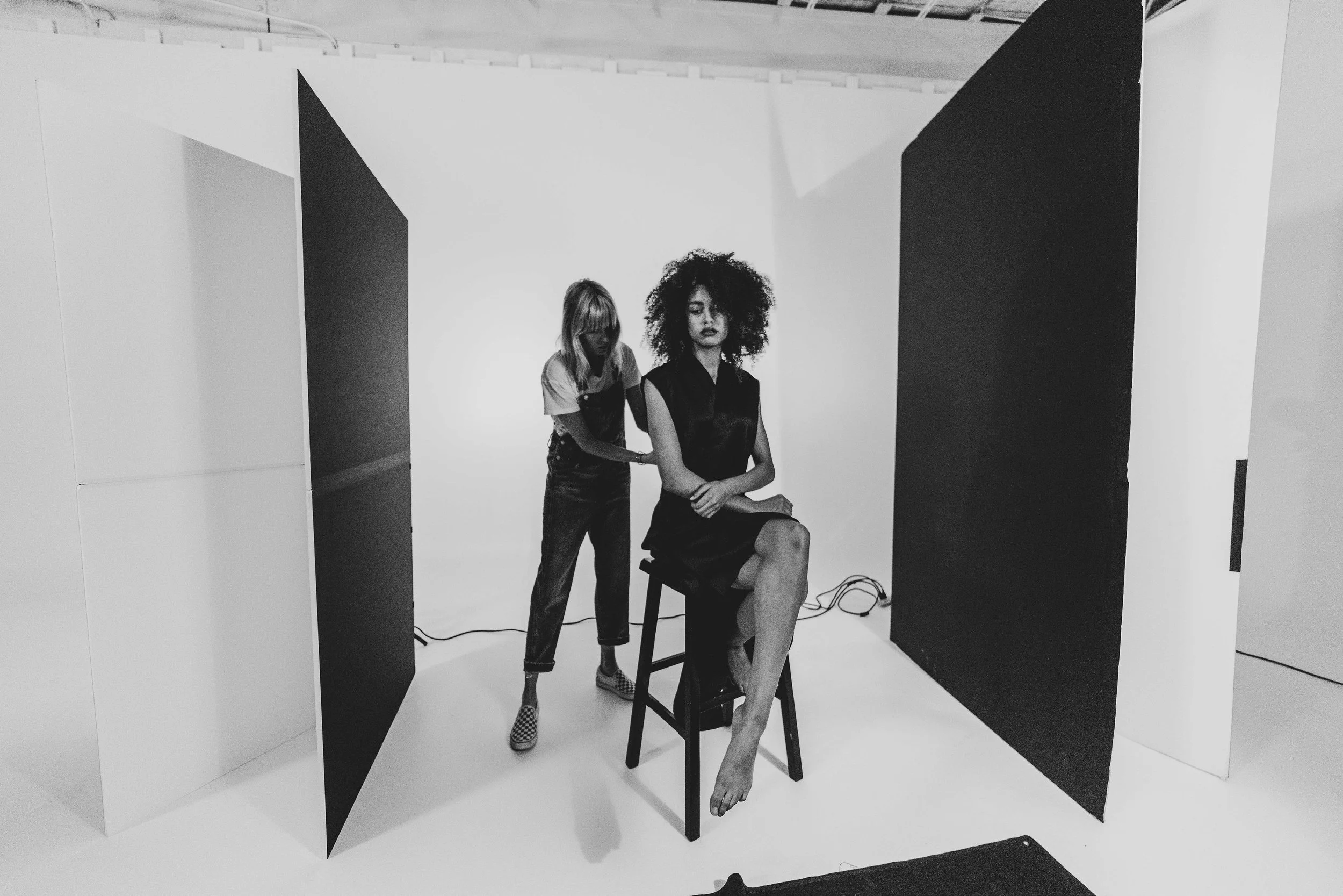 A woman with curly hair sitting on a stool in a photography studio, with her legs crossed and her hands resting on her lap, while another woman with straight hair stands behind her, adjusting her dress. The studio has a plain background with black and white elements, and there are cables on the floor.