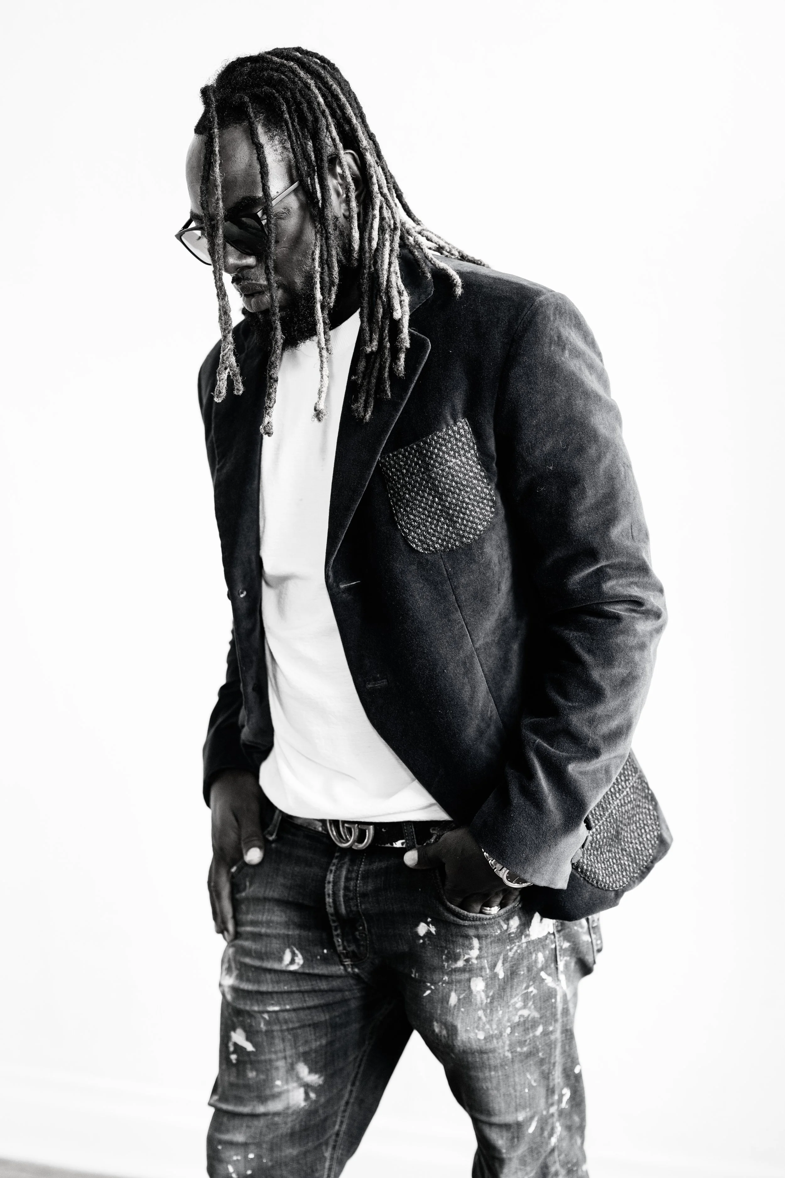 Black and white photo of a man with dreadlocks wearing sunglasses, a blazer with a patterned pocket, a white shirt, and paint-stained jeans.