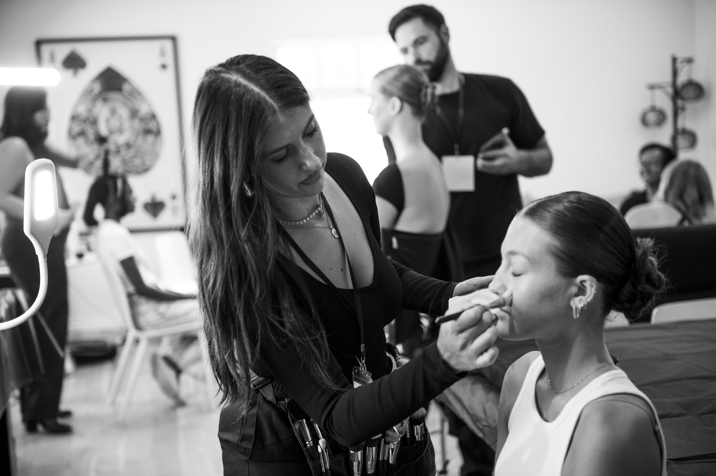A woman with long dark hair applying makeup to a seated young woman at a beauty event.