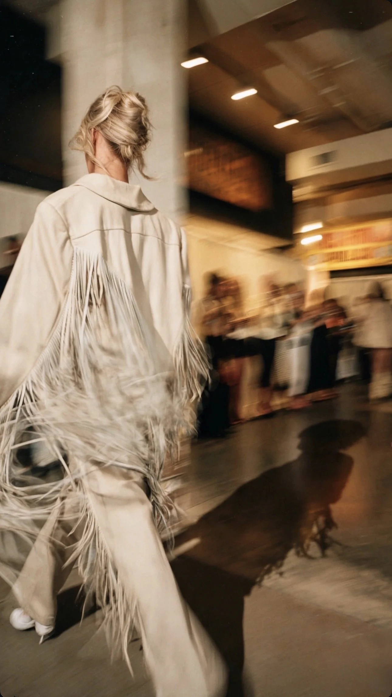 Woman with blonde hair styled in loose waves, wearing a cream-colored fringed jacket, walking in an indoor venue with blurred background of people and warm lighting.