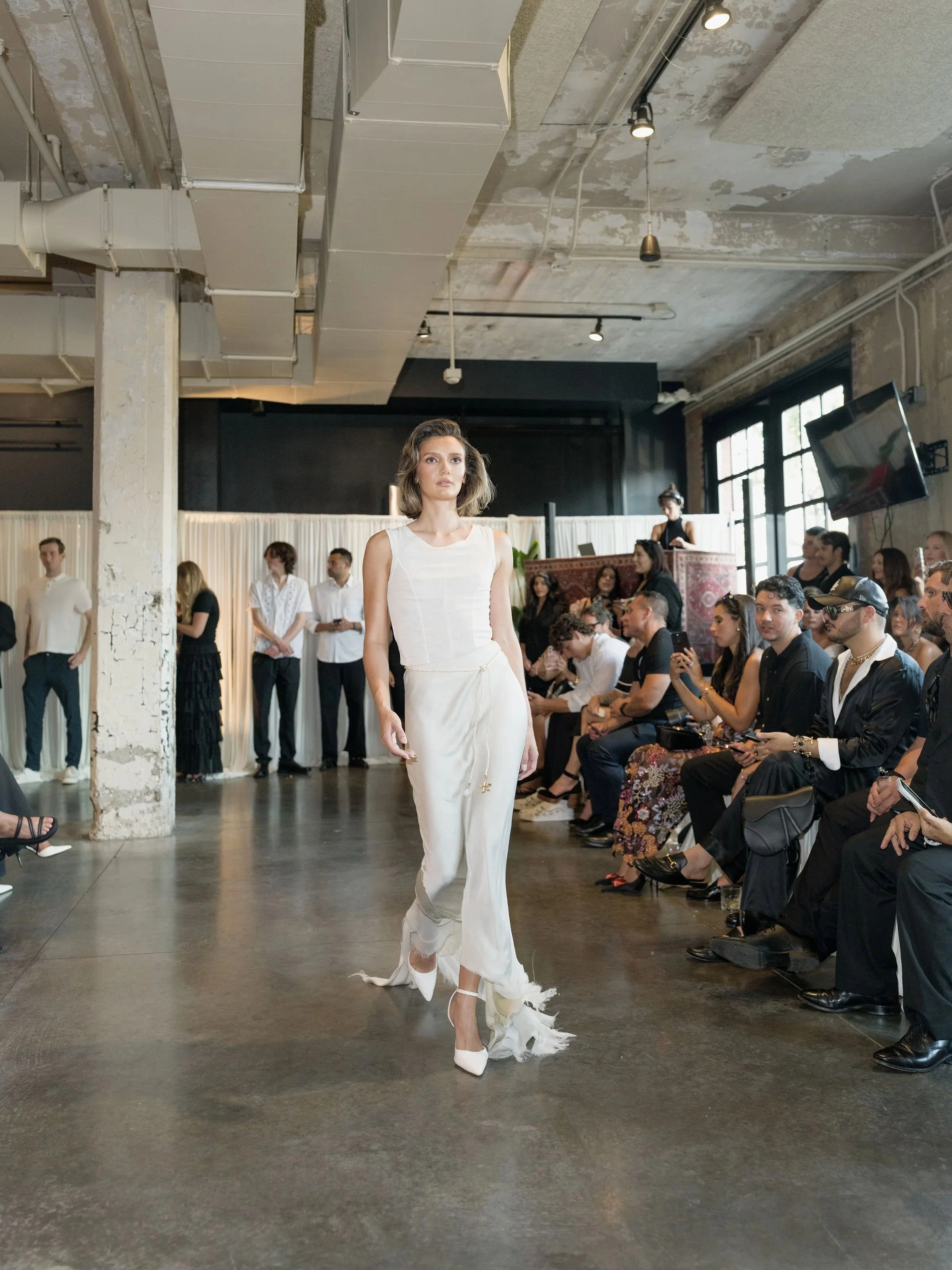 A fashion runway show indoors with a female model walking on the runway. The model is wearing a white sleeveless top and wide-legged white pants with decorative fringes. Audience members are seated on the right side, watching the show, and some are taking photos. The industrial interior features exposed concrete, large windows, and ceiling ducts.