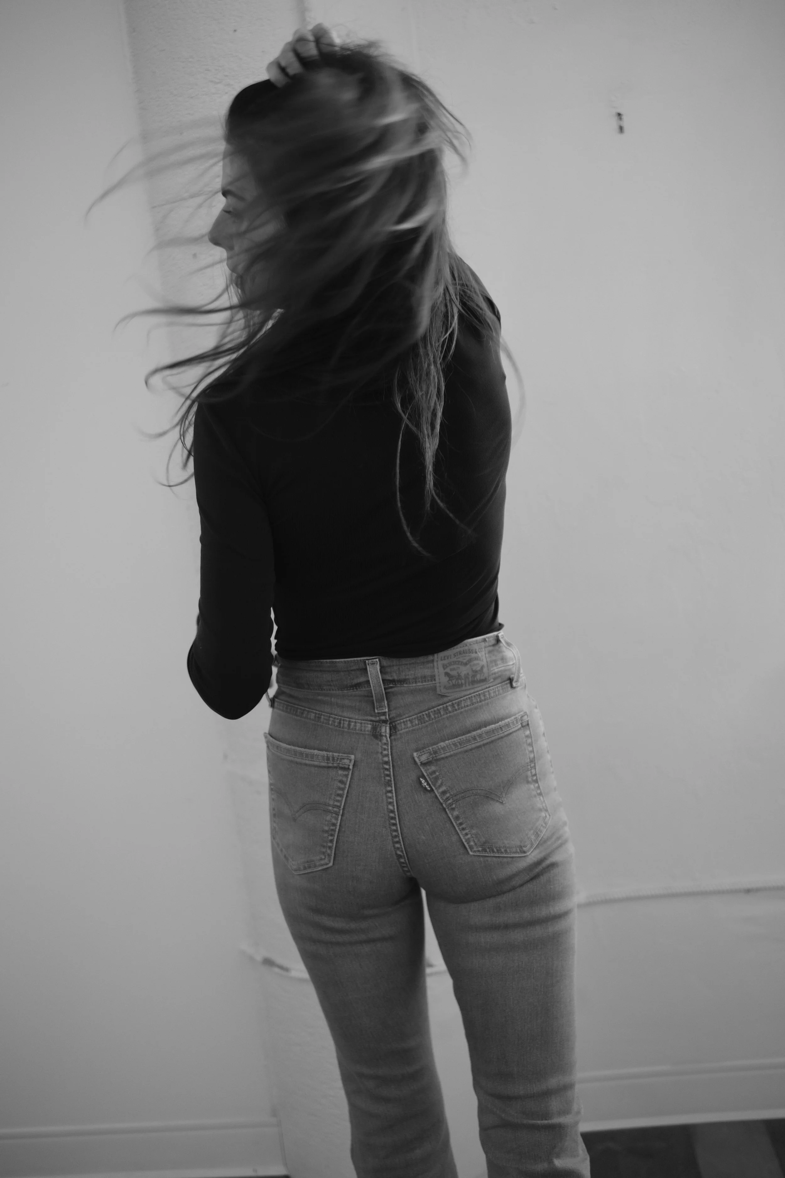 Black and white photo of a woman with long hair, wearing a black top and jeans, standing indoors with her back to the camera, her head turned to the side and her right hand on her head.