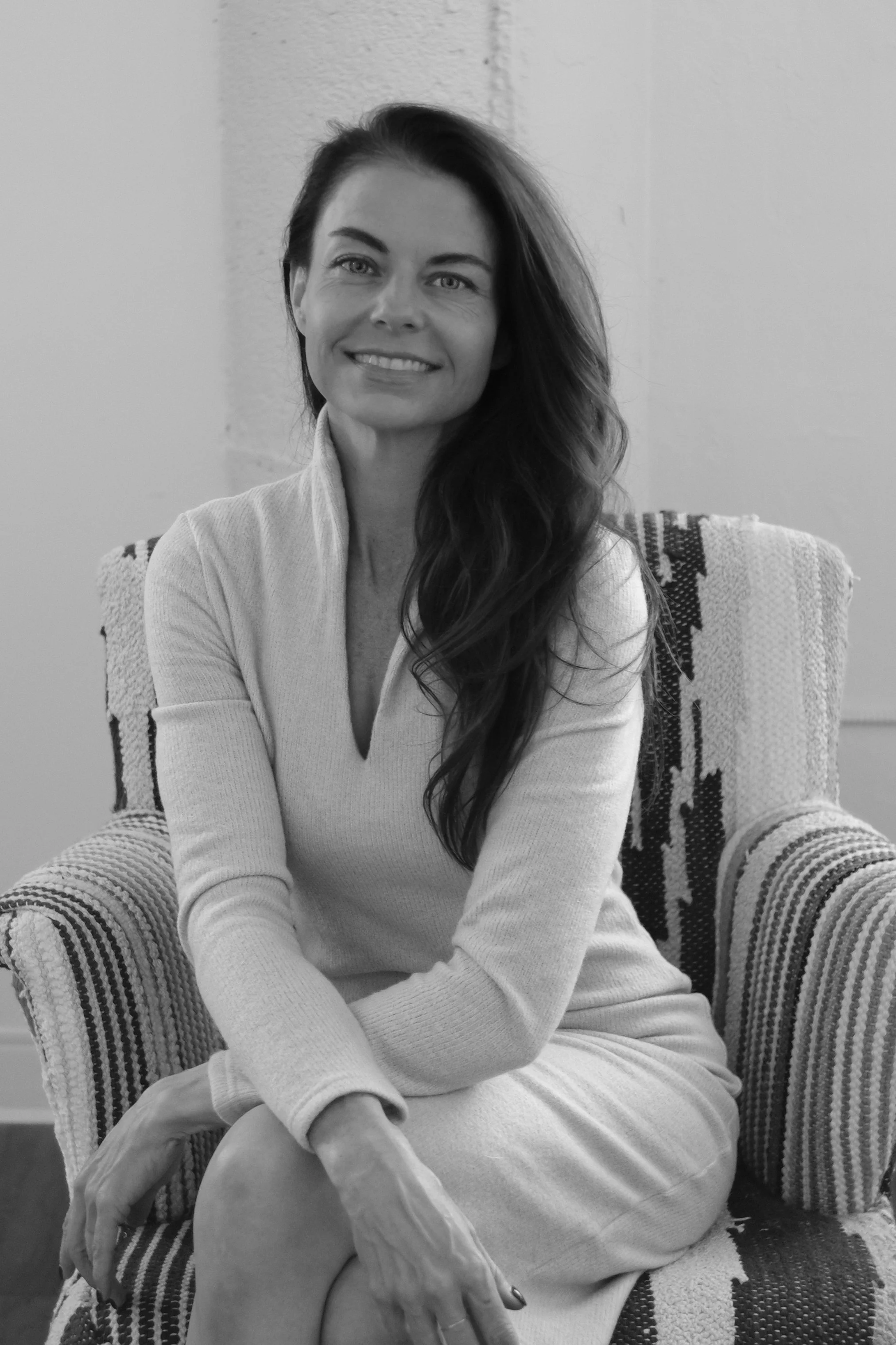 A smiling woman with long, wavy hair sitting cross-legged on a patterned armchair, wearing a light-colored sweater and skirt, in a room with a plain wall.