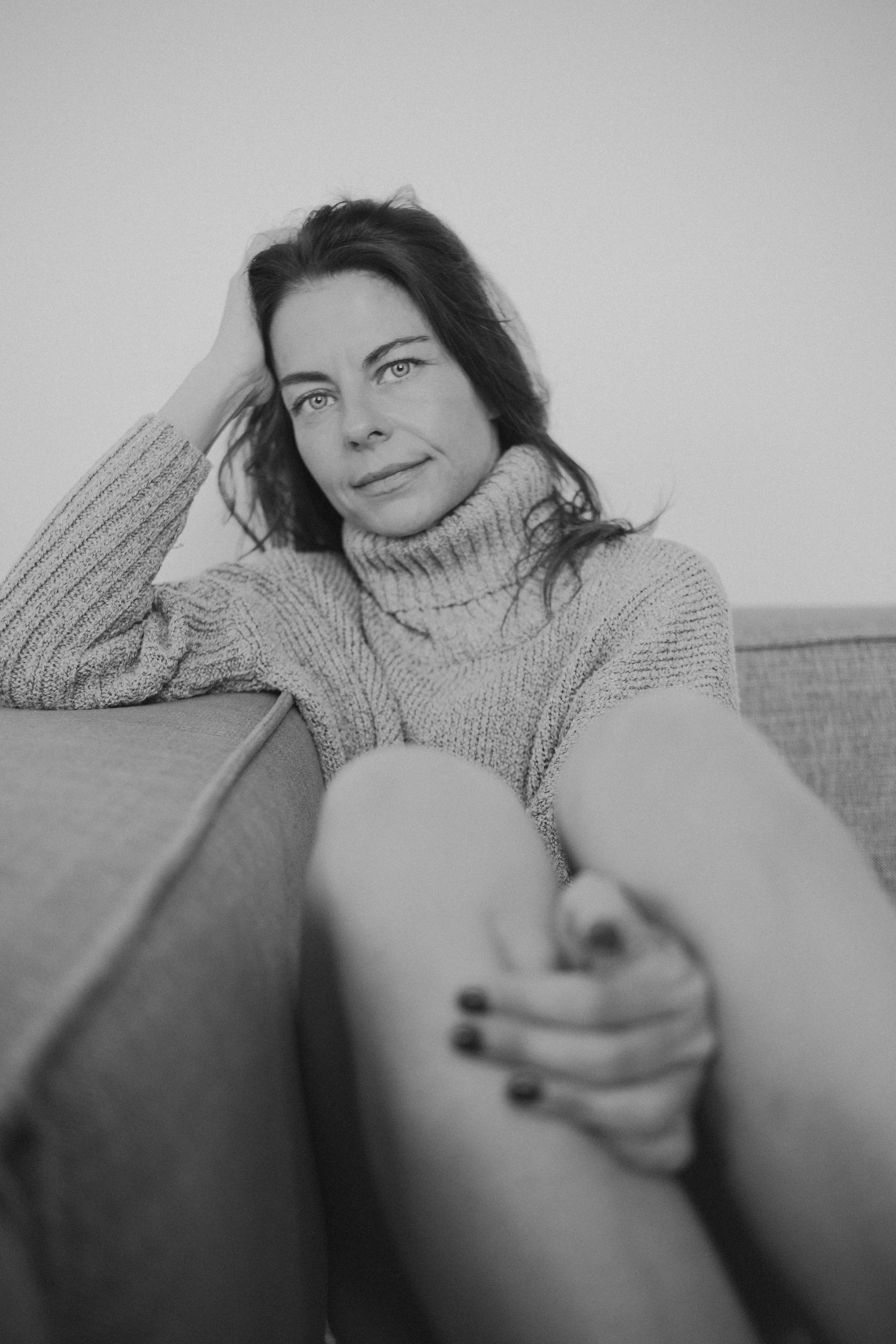 A woman with dark hair and light-colored eyes, wearing a turtleneck sweater, taking a selfie while reclining on a couch with plain walls in the background. The photo appears to be taken in a relaxed, casual setting.