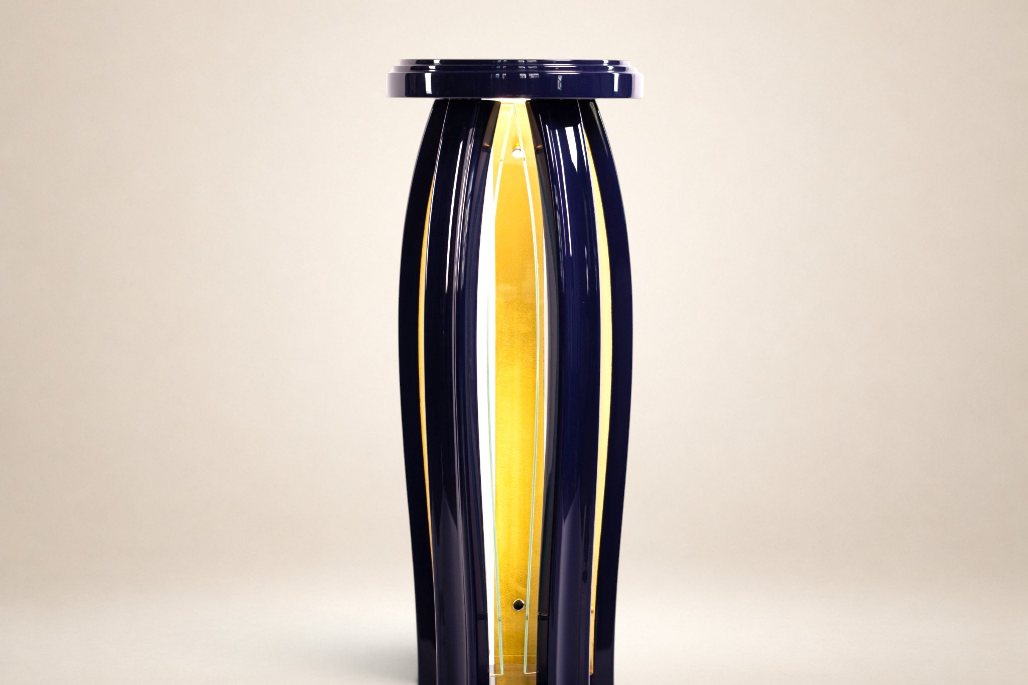 A tall, modern table lamp with a unique design, featuring multiple dark blue, curved, overlapping panels and a vertical strip of warm yellow light in the center, against a plain, off-white background.