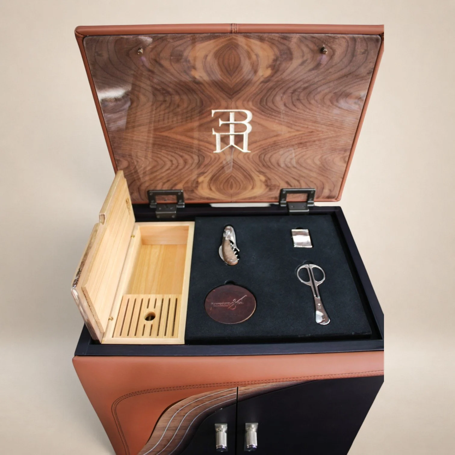 A wooden jewelry or watch box with a black velvet interior. It contains a pair of scissors, a round object, and a small metallic item. The box's interior lid has a wood grain pattern with a white logo in the center.