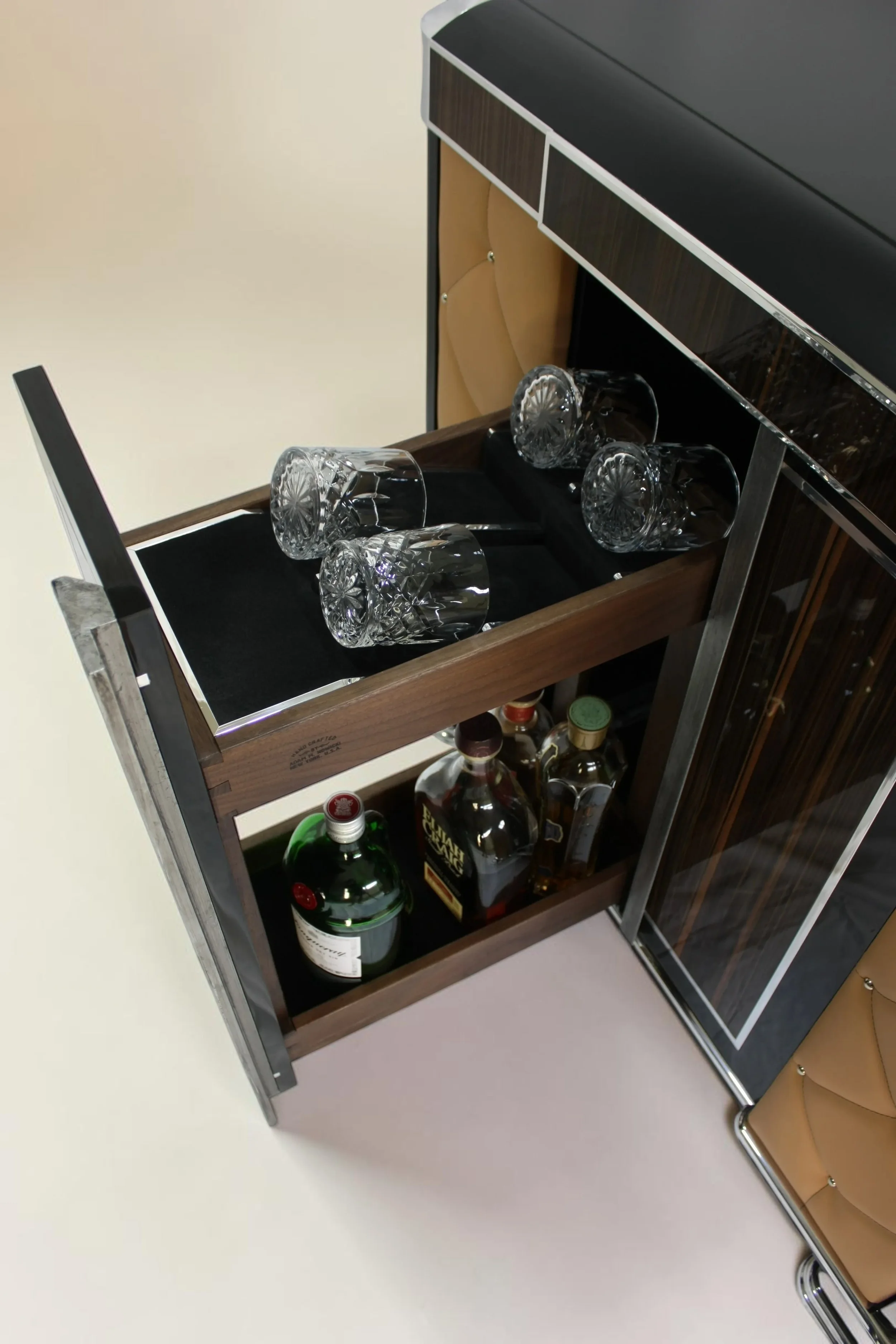 Bar cart with four empty glasses on top, containing a black cloth, and bottles of alcohol on a lower shelf, including a green gin bottle, whiskey, and bourbon. The bar cart is in a room with beige walls and a brown leather chair with a metal frame ne