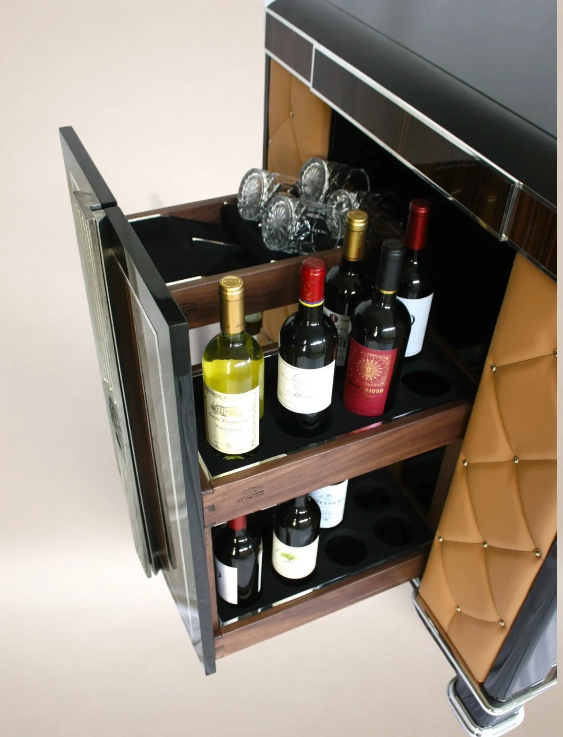 A wooden bar cart with bottles of wine, wine glasses, and a wine bucket, with a tan cushioned chair beside it.