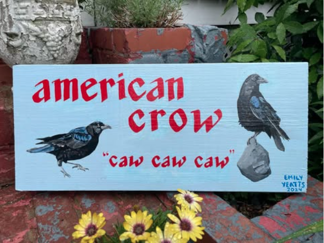 A painted wooden sign features the words 'American Crow' in red, with the phrase 'caw caw caw'. The sign has illustrations of two American crows. It is placed outdoors among plants and flowers, signed by Emily Yeatts in 2024.