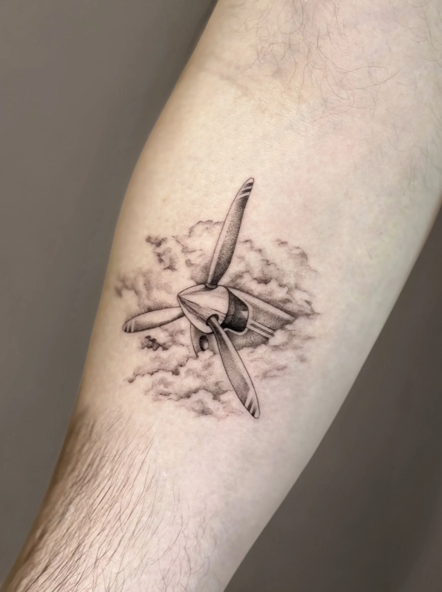 Black and grey fine line plane tattoo with detailed propeller and fine line shading, surrounded by abstract clouds