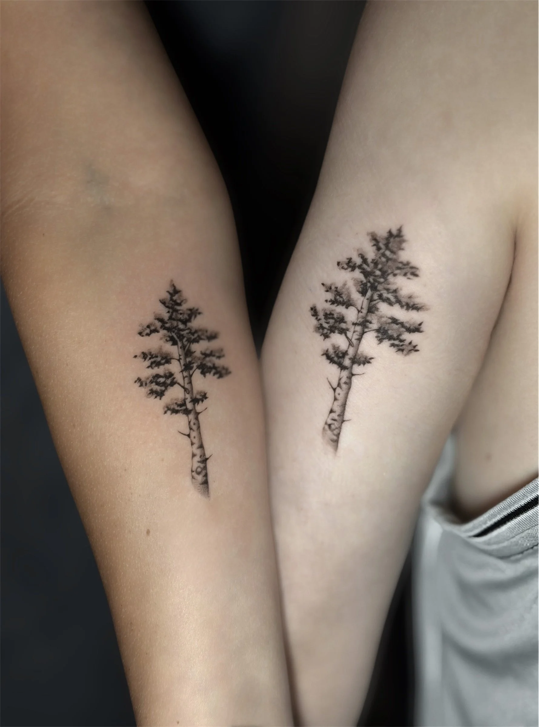 Matching black and grey tree tattoos on two arms, featuring delicate branches, detailed leaves, fine line work, and soft shading.