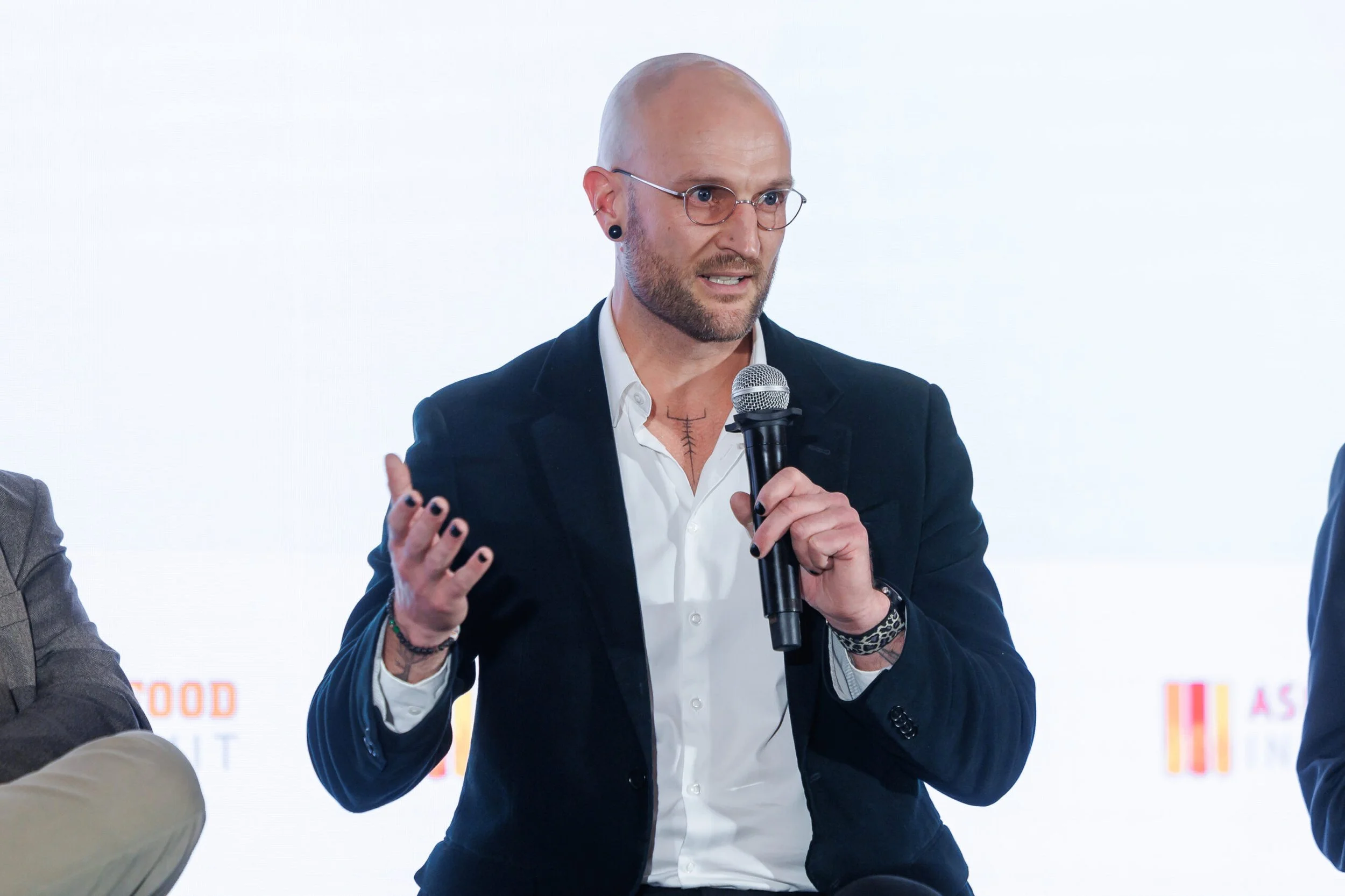 A man in a dark suit, white shirt, with glasses and tattoos visible on his neck and arms, speaking into a microphone during an event.
