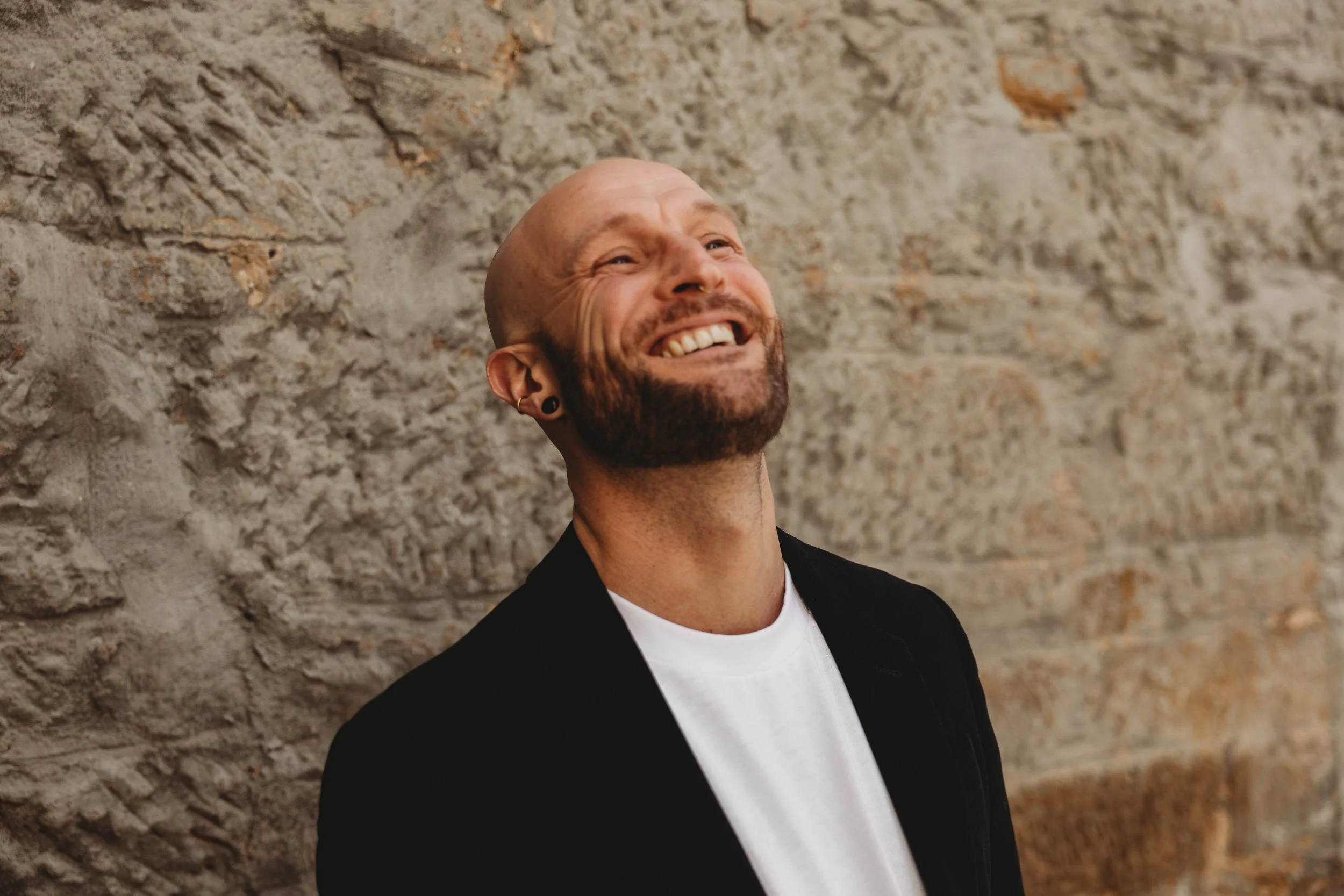 A smiling man with a beard, hoop earrings, and a towel, standing against a stone wall.